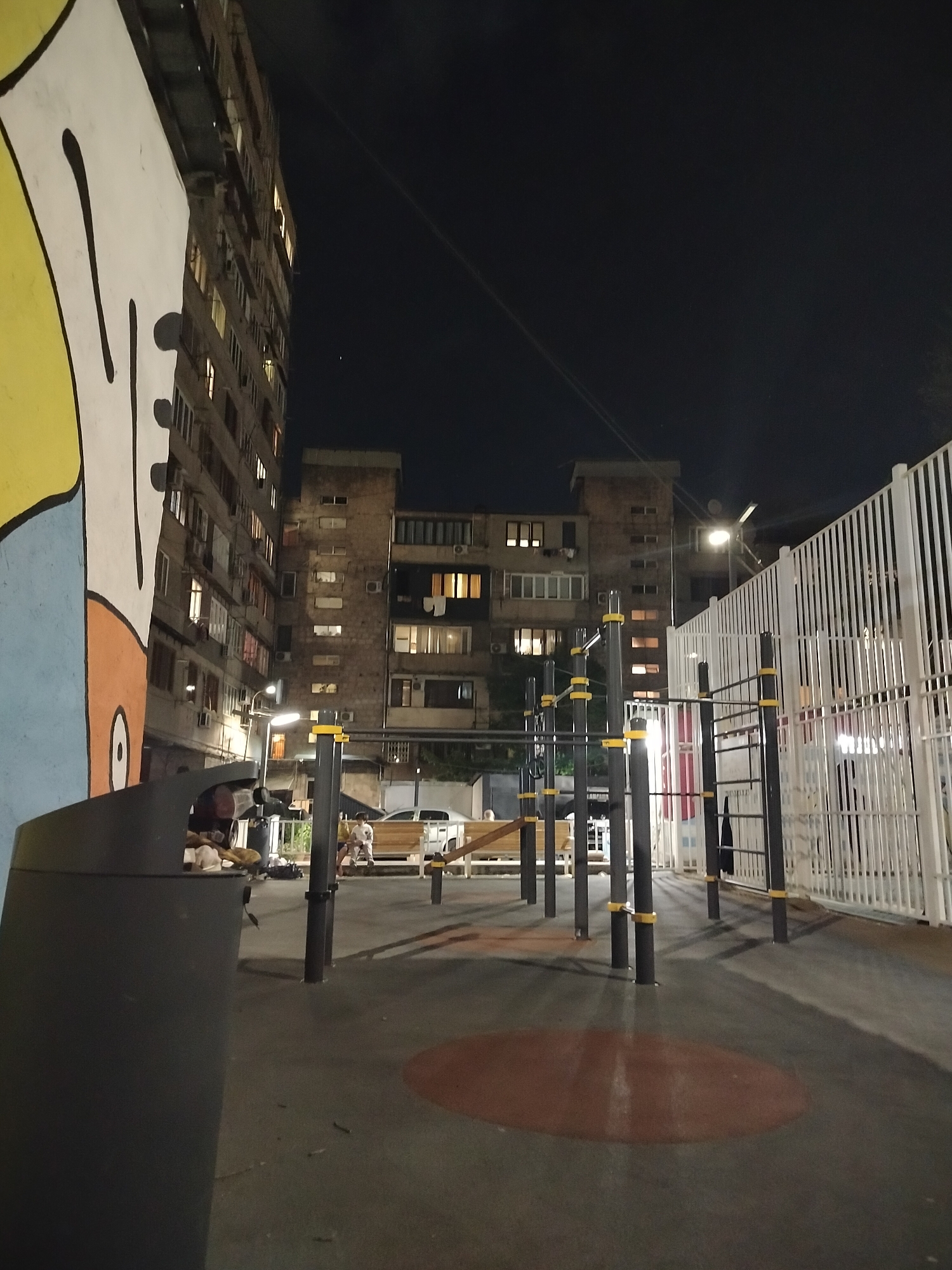 The image shows a playground at night in Yerevan, Armenia. The playground includes pull-up bars with yellow accents, situated on a gray surface with a red circular design. A white fence borders the playground. In the background are apartment buildings with lit windows. A street light provides illumination. A trash can is visible in the foreground, partially obscuring a colorful mural. In the background, some individuals are seated on a bench. A white vehicle is parked nearby.