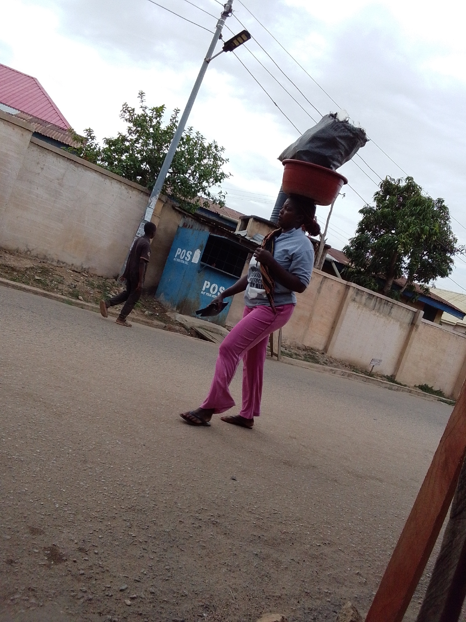 An outdoor, slightly low-angle shot captures a bustling street scene in Jos, Nigeria, under an overcast sky. The primary subject is a woman walking from left to right, skillfully balancing a red basin, topped with a large dark bag and what appears to be ice, on her head. She wears a light-colored short-sleeved top with white text on the front and bright pink pants, accessorized with sandals and a fabric draped over her left shoulder.
In the mid-ground, a man in dark clothing walks away from the camera, moving towards the left side of the frame. Behind him are two distinct blue kiosks or small structures. Both kiosks feature the white text "POS" (Point of Sale) prominently, with additional, partially visible text below that reads "PAY YOUR HERE" (likely "PAY YOUR BILL HERE"). A tall utility pole, fitted with a street light and multiple overhead power lines, stands between the two individuals.
The background consists of various buildings with corrugated metal roofs, some painted pink, and others darker. Concrete walls separate the properties from the road, with green trees visible above and behind them. The road surface is a textured asphalt. The overall atmosphere suggests a typical daytime urban or semi-urban setting, with no direct sunlight due to the cloudy conditions.