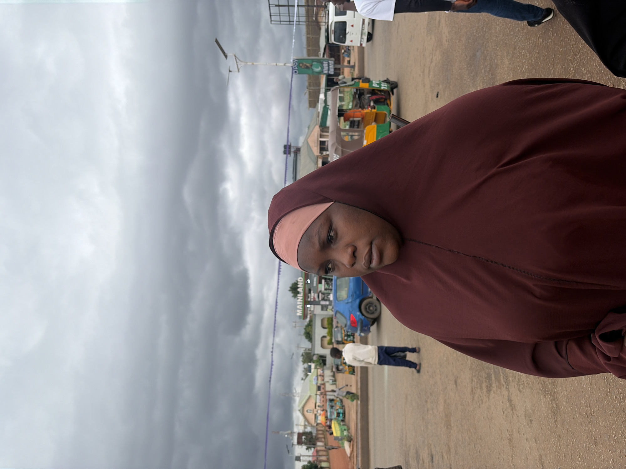 A medium-shot, eye-level, portrait photograph captures a woman in a maroon hijab from the chest up, facing right and looking down and to the side with a neutral expression. Her hijab is a deep maroon color with a lighter pink fabric visible around her forehead. The background, blurred due to the shallow depth of field, depicts a street scene in Gura Top, Nigeria, under a cloudy sky.
In the mid-ground, a blue car is parked on the right side of the street. Behind it, a green and yellow auto-rickshaw is visible, with a person in an orange shirt seated in the driver's position. Several other auto-rickshaws and what appear to be small shops or stalls are further in the background, along with a white van and a white building with a red roof. A sign reading "MAINLAND" is prominently displayed above a gas station. In the middle distance, a young boy in a light-colored jacket and dark pants walks away from the camera. Several power lines are strung across the scene. The ground is a mix of asphalt and dirt.