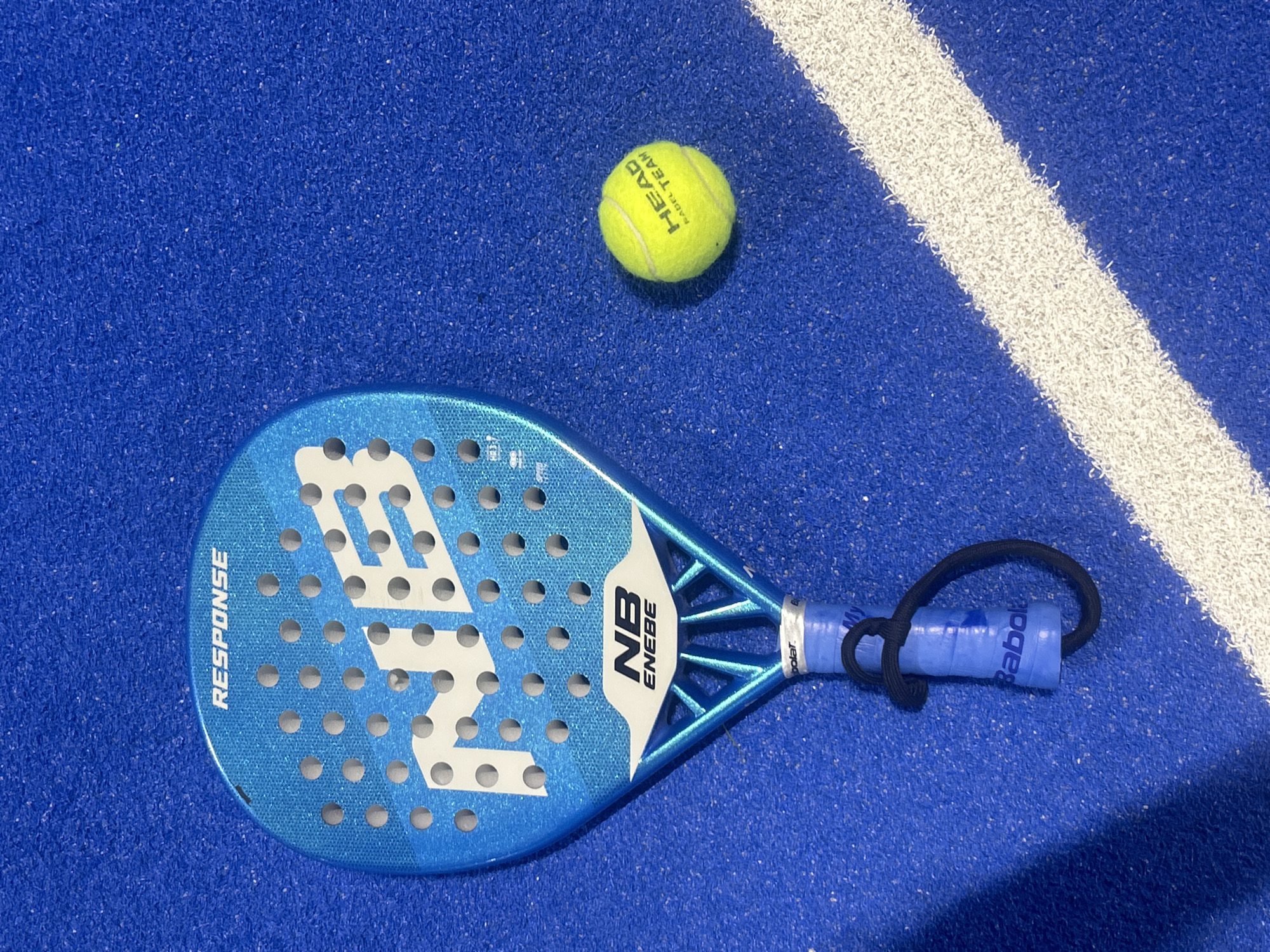 A blue paddle racket with white lettering and a blue handle is lying on a blue surface with a yellow tennis ball nearby. A white line is also visible on the blue surface. Based on the court markings and equipment, the image appears to be a padel tennis court. The location in the city of Pleven, Bulgaria, is not discernible from the image.