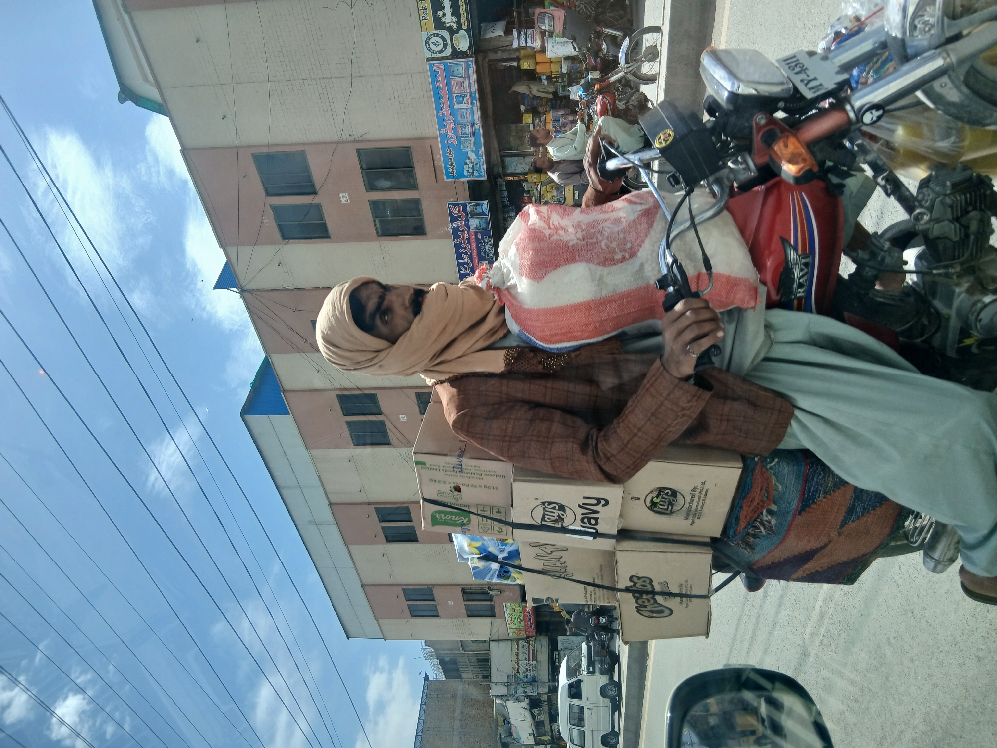 An outdoor shot captures a man on a heavily loaded motorcycle, likely transporting goods, in what appears to be an urban setting. The man, seen from the chest up, wears a light brown head covering or turban, a brown checkered jacket, and light blue/grey trousers. He has a beard and is holding the motorcycle handlebars.
The red motorcycle, branded "RAVI" on its fuel tank, is laden with multiple cardboard boxes and a large white and red striped sack. Visible text on the boxes includes "Knorr Instant" and "Lay's Javy," with one box also stating "Manufactured By: 31.5g x 72 Packs = 2.2 kg Unilever Pakistan Limited." A patterned rug or textile is also draped over part of the motorcycle. The license plate "MY-4311" is visible on the motorcycle's front.
In the background, two men are visible near a storefront. The street is lined with multi-story buildings, some with multiple windows and balconies. Several business signs, predominantly in Urdu/Arabic script, are visible, including "الحکمت ٹریڈرز مولا سیلانین" (Al-Hikmat Traders Maule Sailaniyan), "صالح محمد اینڈ" (Saleh Muhammad &), "گل فرید رز مل کل" (Gul Fareed Rice Mill), and one with "Park" and a coffee cup logo. Electrical wires crisscross the bright blue sky, which has scattered white clouds. The scene suggests a busy daytime environment, typical of a market or commercial street. Other vehicles, including a white van or mini-truck and additional motorcycles, can be seen in the distance on the road.