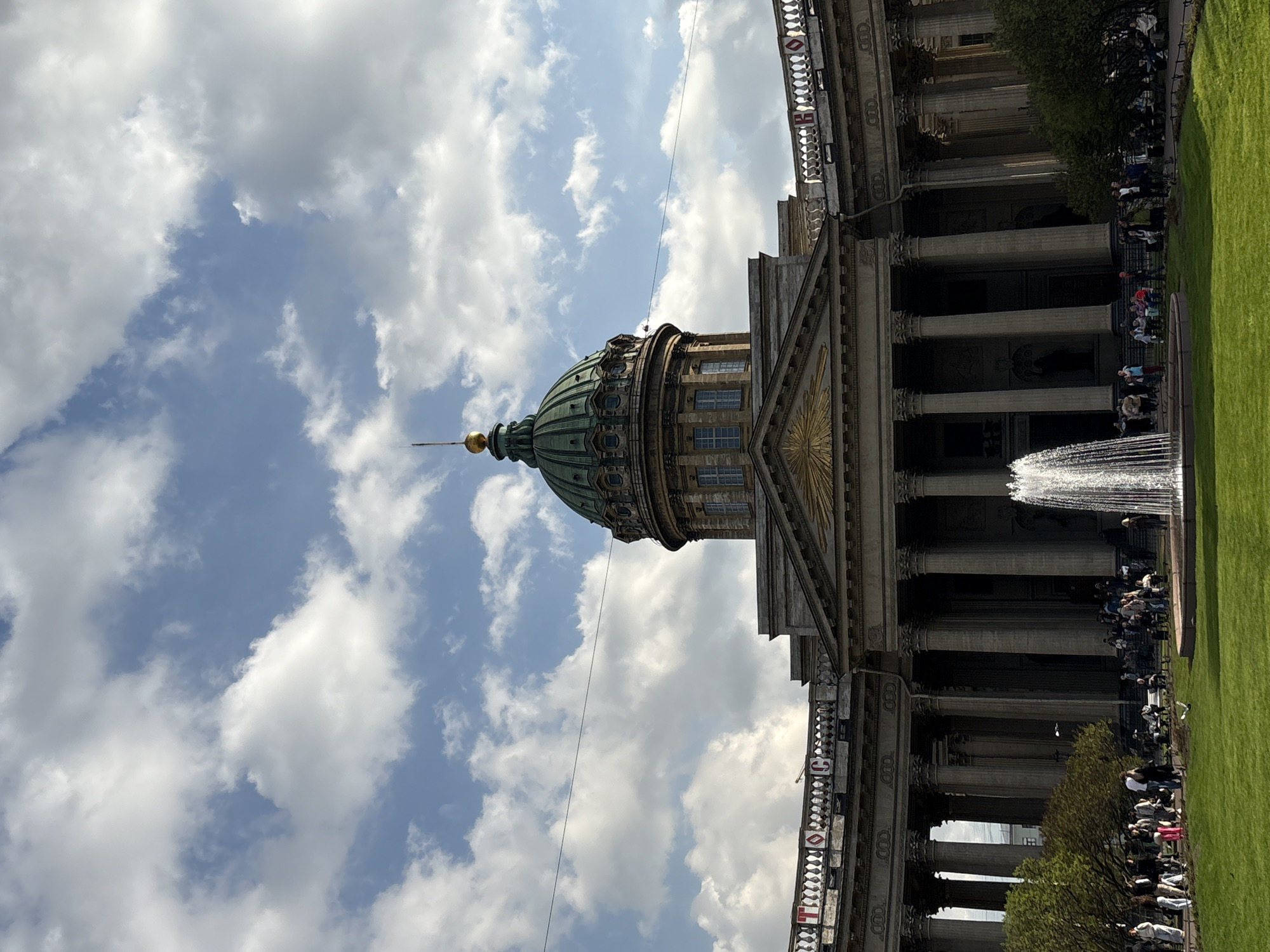 The image depicts the Kazan Cathedral in Saint Petersburg, Russia. The cathedral's distinctive green dome is visible against a backdrop of a cloudy sky. The cathedral's facade features classical columns and architectural details. A fountain is in the foreground. People are present near the building, presumably visiting or sightseeing. The photograph is taken from a low angle, emphasizing the height and grandeur of the cathedral. A patch of green grass suggests a lawn or park setting.