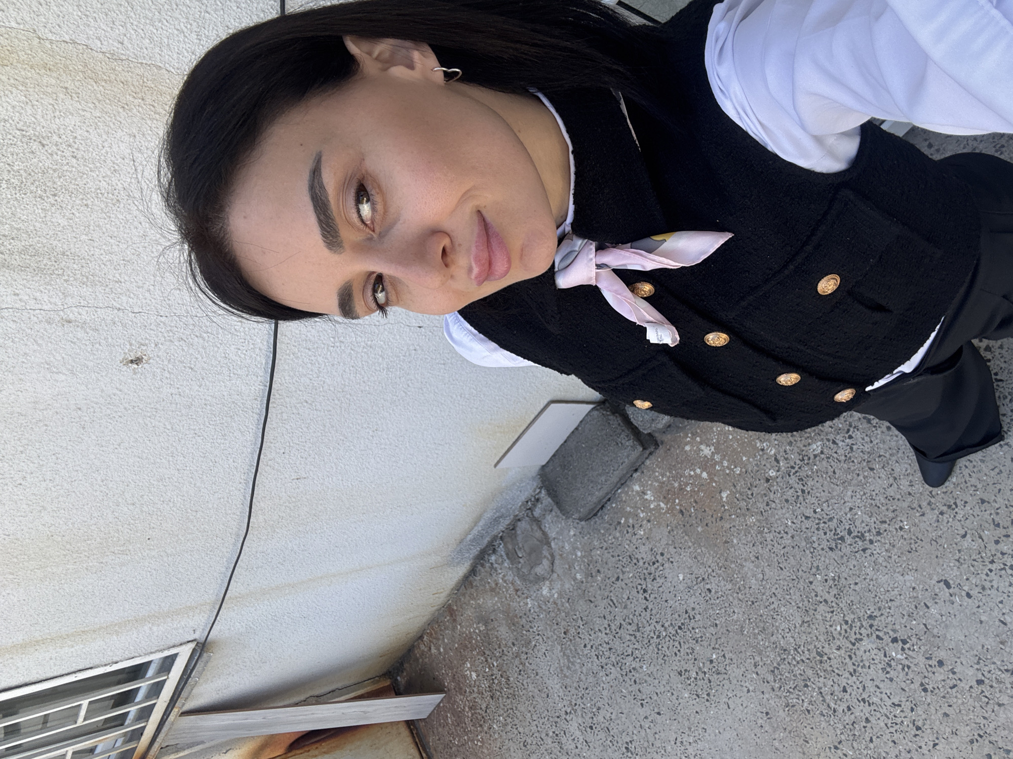 A high-angle, medium shot captures a young woman outdoors, looking directly at the camera with a subtle smirk. She has dark hair, dark eyebrows, and fair skin. She is dressed in a white collared shirt, layered with a black textured vest featuring several gold buttons. A light pink scarf with a printed pattern and faint, partially illegible text (appearing to include "CERAMIC MADE IN") is tied around her neck. A small heart-shaped earring is visible in her left ear.
The setting appears to be an urban exterior. To the left, a light-colored, textured wall (possibly stucco or painted concrete) shows some minor cracks and discoloration. The ground is a dark, speckled concrete surface. At the bottom left, a metal grate, possibly a window or vent, is partially covered by some wooden planks. A concrete block and a small white tile are also visible near the base of the wall. The bright, even lighting suggests it is daytime. No specific activity beyond posing for the photograph is depicted.
