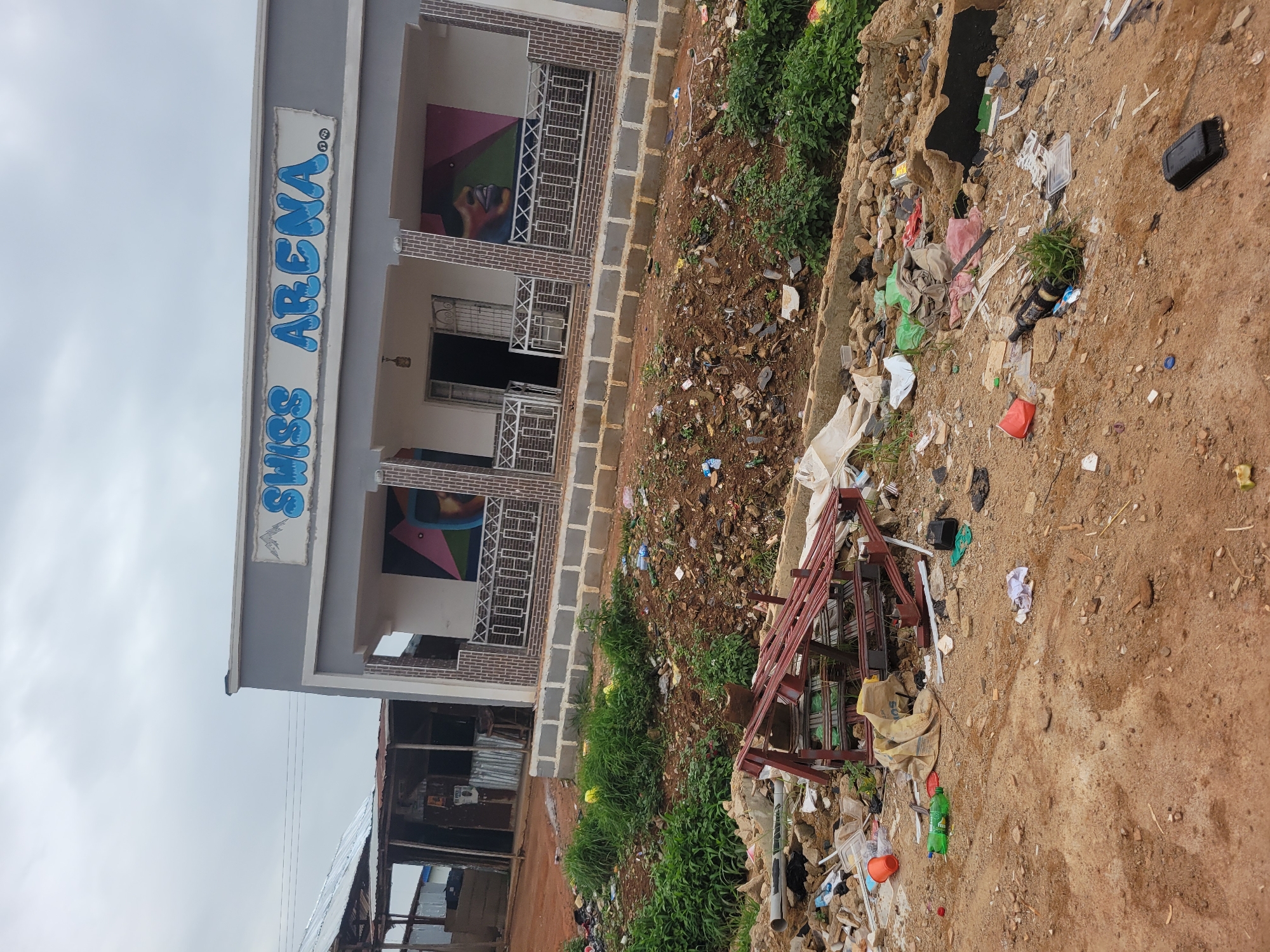 The image depicts a scene of significant litter and debris in front of a building with the text "SWISS ARENA" visible in blue lettering on its facade. The building appears to be located in Durumi, Nigeria, based on the provided context. The foreground and midground are heavily strewn with various types of trash, including plastic bottles, bags, packaging, construction debris, and what appears to be a discarded metal frame structure. Vegetation, primarily grass and some bushes, is interspersed within the debris. The building itself has a light gray exterior with decorative brickwork around the entrance and on the balcony. There are large artistic murals on the exterior walls of the building, depicting abstract shapes and a stylized face. The sky is overcast. Adjacent to the main building, there is a smaller, more dilapidated structure with a corrugated metal roof, also appearing to have debris around it. The overall impression is one of neglect and poor waste management in the surrounding environment.