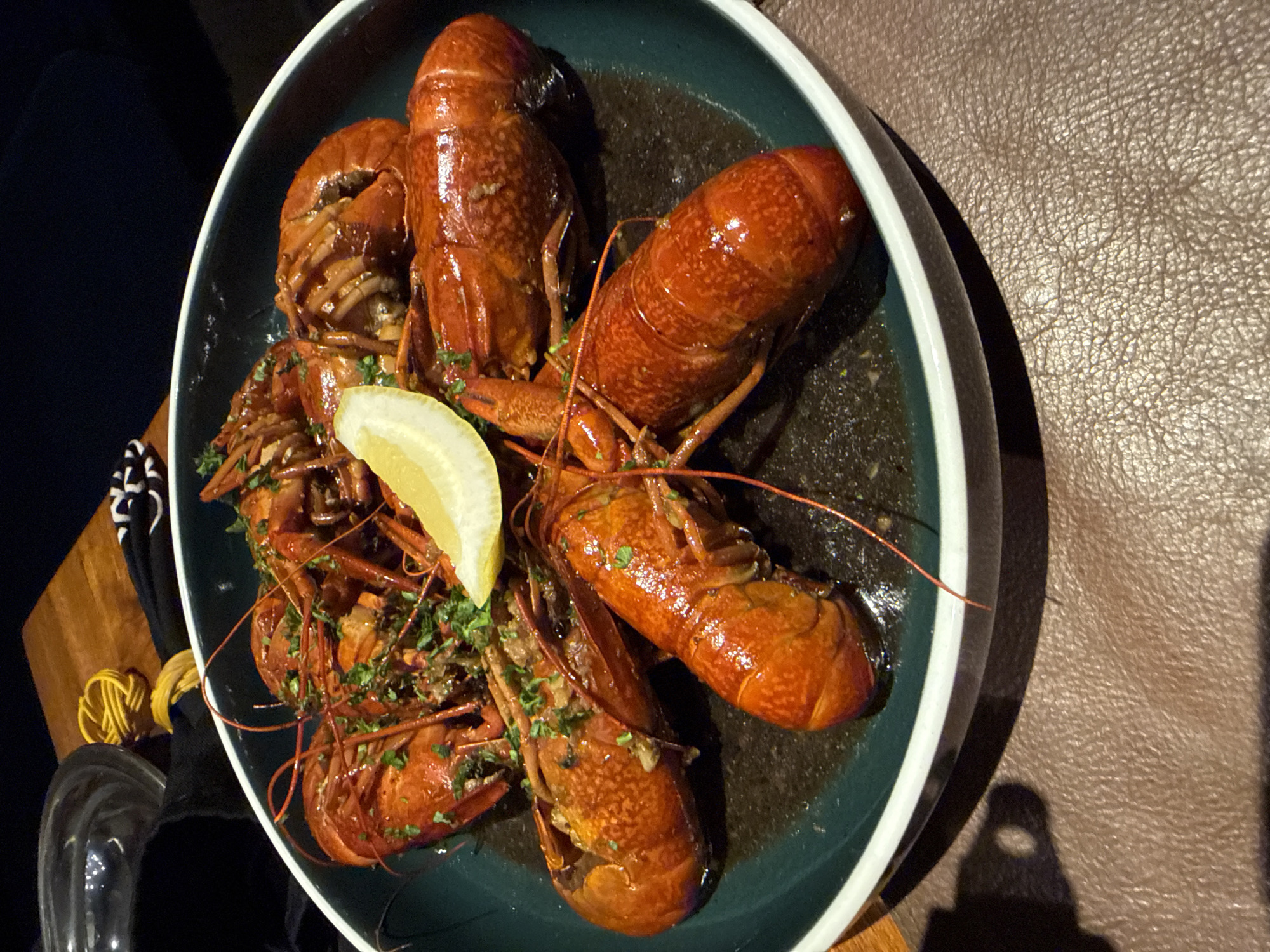 A close-up, top-down shot captures a serving of cooked crustaceans, likely crayfish or small lobsters, arranged on a dark green plate with a white rim. There are at least six crustaceans visible, characterized by their bright reddish-orange shells, some appearing split open to reveal meat. They are generously garnished with chopped green herbs, possibly parsley or cilantro, and a single lemon wedge rests in the center of the plate, adding a bright contrast. The crustaceans are resting in a dark liquid or sauce within the plate.
The plate is positioned on a dark brown, textured surface that resembles leather or a similar material. To the left of the plate, a dark cloth or napkin is visible, partially covering a small wooden cutting board. On the board, there are a few coiled yellow items, possibly small strings or elastic bands. A clear glass or container is also partially visible at the bottom left. The background beyond the immediate table area is dark, suggesting an indoor setting with focused lighting on the dish. No people or visible text are present in the image. The scene depicts a meal prepared for consumption. The location context provided is Kuta, Indonesia.