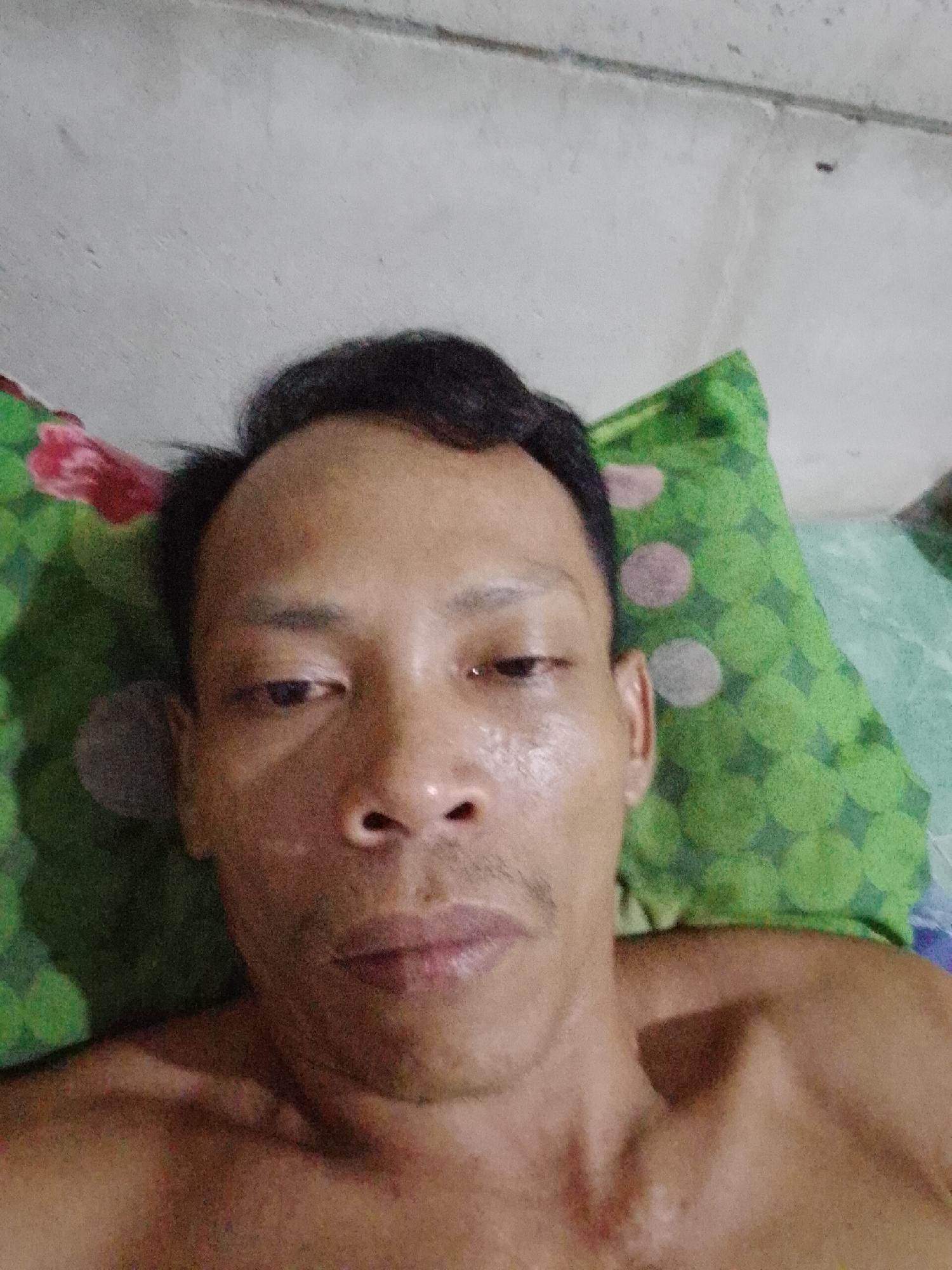 A close-up selfie of a shirtless man, seemingly lying down on a patterned pillow and bed. His face is moist with visible pores. The background features a wall, possibly concrete. The image was taken in Bukit Ketok, Indonesia.