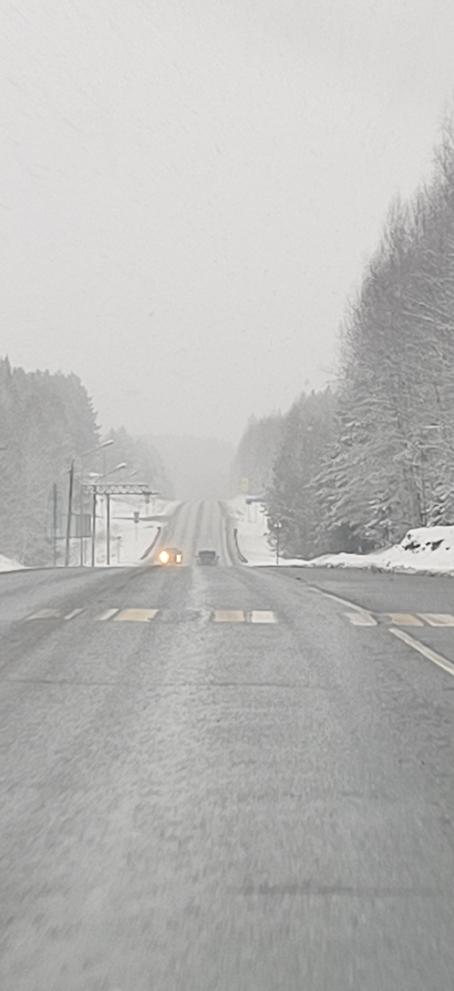 The image depicts a snowy, overcast day on a road outside Chaykovsky, Russia. The road stretches toward the horizon, where visibility is significantly reduced by the snowfall. Trees, heavily laden with snow, line either side of the road. There are two vehicles on the road ahead: one closer, with headlights visible, and another further away. Yellow traffic-calming strips are visible on the road surface. Streetlights and power lines are visible on the left side of the road.