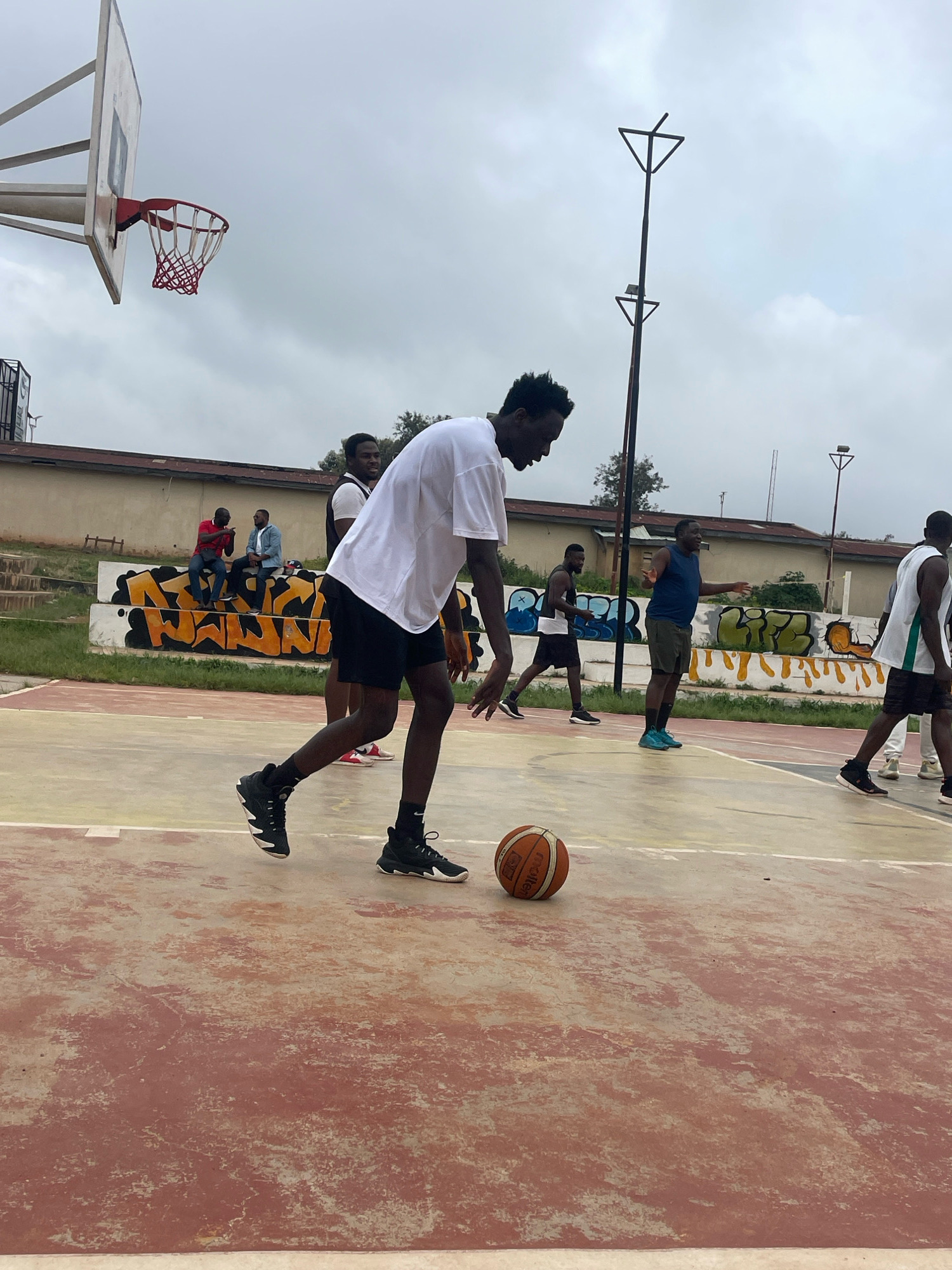 The image shows a basketball court in Gura Top, Nigeria. The court is made of concrete and is weathered. There are several people on the court playing basketball. In the foreground, a young man in a white t-shirt and black shorts is dribbling a basketball. In the background, other people are playing or watching. There is a basketball hoop in the upper left corner of the image, and a fence in the background. The sky is cloudy.