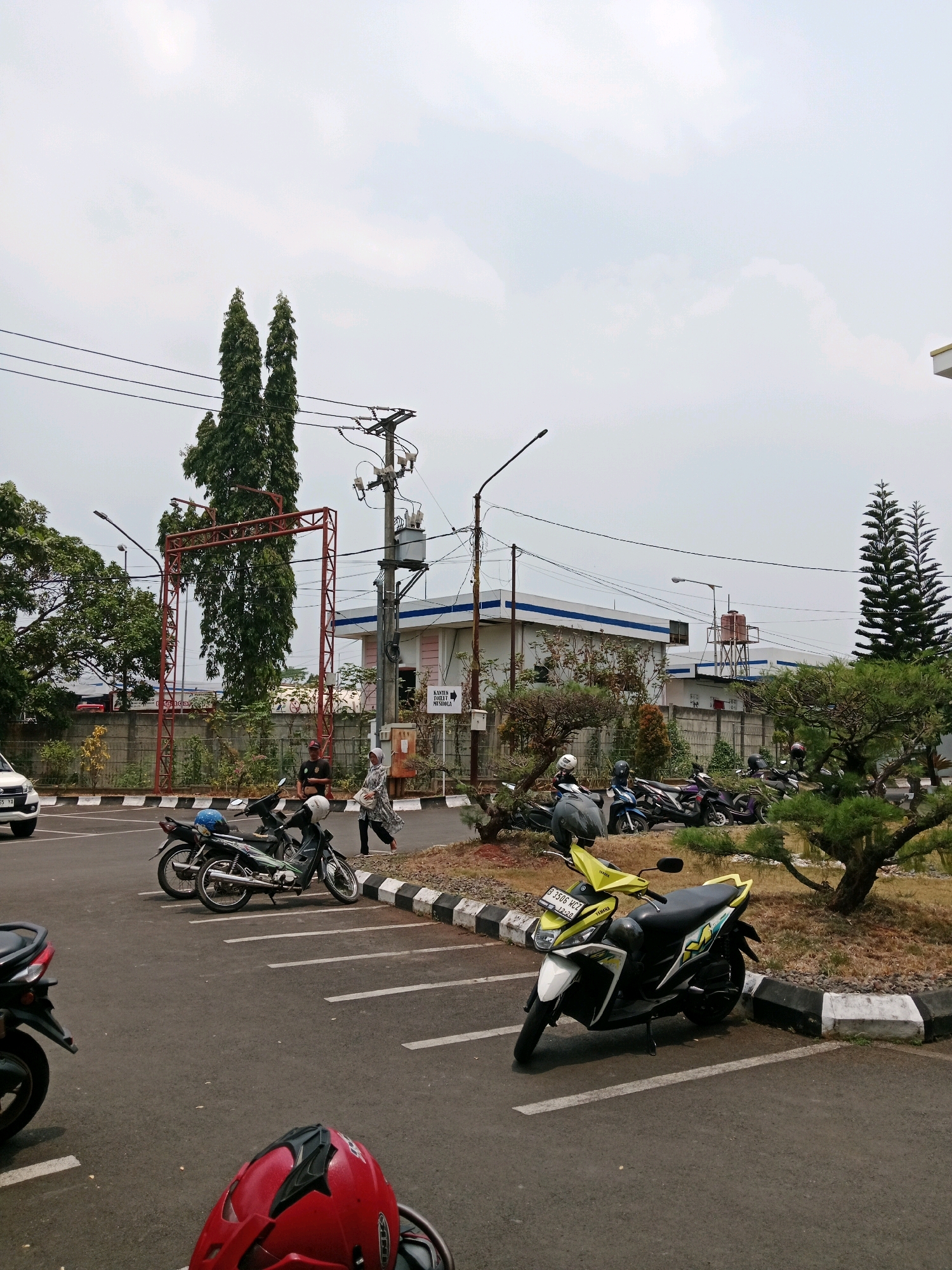 The image depicts an outdoor scene in Karangtengah, Indonesia. In the foreground, a red helmet is placed on what appears to be a motorcycle. The middle ground features a parking lot with several parked motorcycles, including a prominent yellow and white one with a license plate "B 3506 HCZ". White parking lines are visible on the asphalt. Several people are walking or standing in the background. One person, dressed in a grey and white patterned outfit and a headscarf, is walking past parked motorcycles. Another person in a dark shirt and hat is standing near a red metal structure. Behind them, a building with white walls and blue trim is visible, along with a water tower. Power lines and a utility pole with a transformer are present in the scene, as well as various trees and shrubs, including two tall, slender trees in the distance. The sky is overcast.