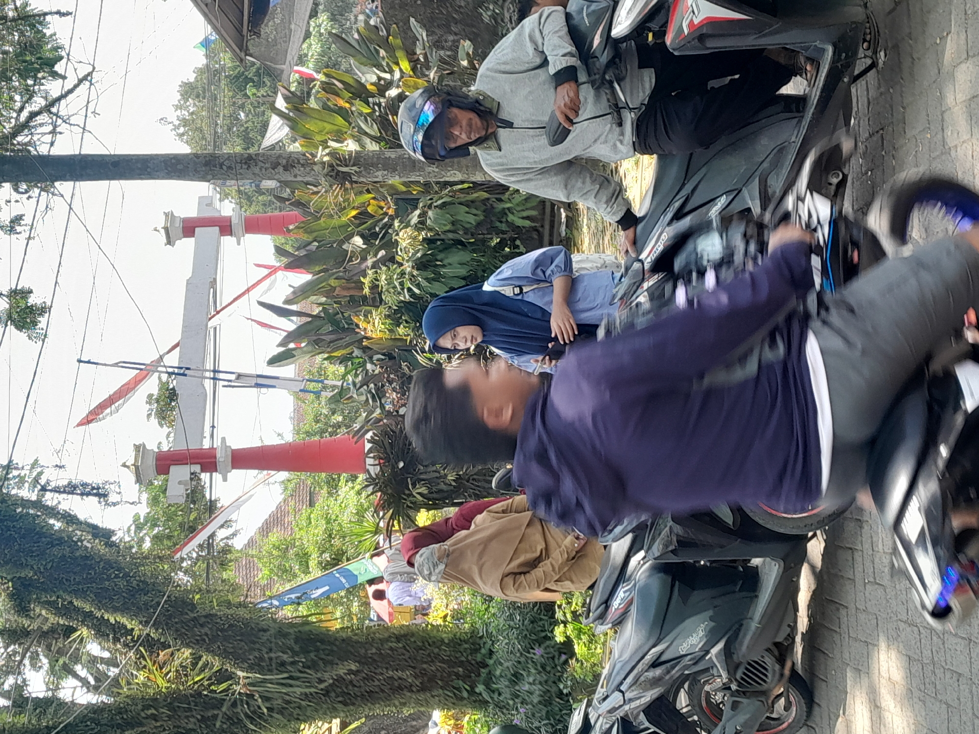 In Genteng, Indonesia, a street scene unfolds with several individuals and motorcycles. A man wearing a gray jacket and helmet sits on a black motorcycle, looking to his right. Next to him, a woman in a blue hijab and light blue shirt is holding a phone. Behind them, two women in hijabs, one in tan and the other in maroon, stand. A man in a purple jacket is in the foreground, blurred due to motion, riding a motorcycle. The background features a red and white gate structure, lush green foliage, and trees with moss. The ground is paved with bricks.