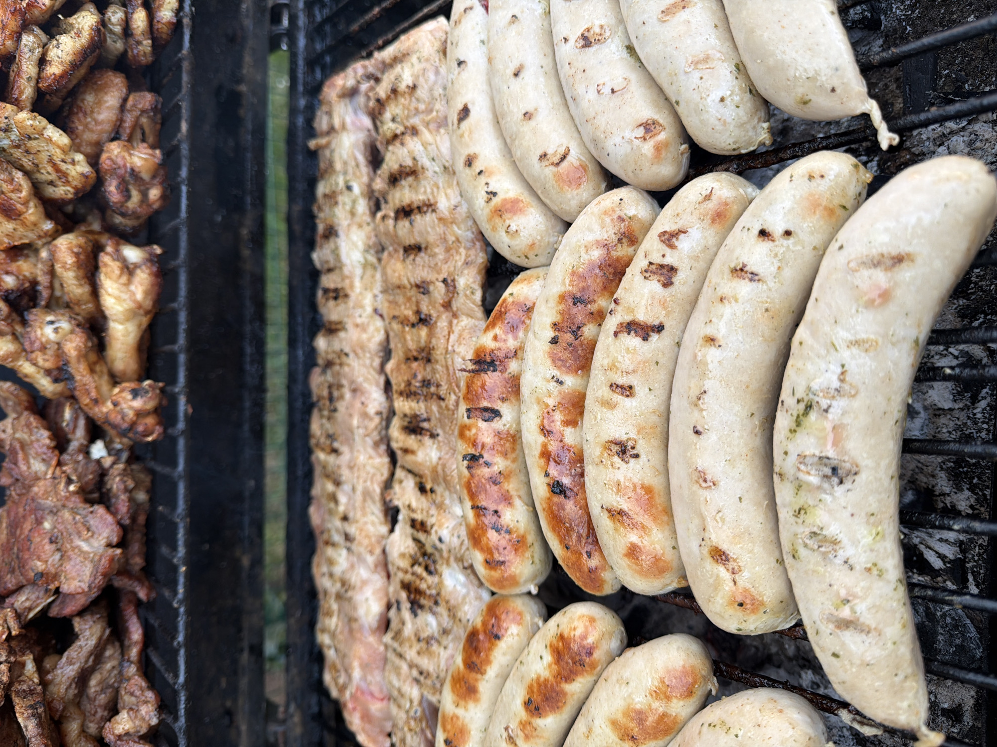 The image shows a grill with various meats cooking on it. On the left, there are pieces of cooked meat, potentially chicken or pork. Next to that, there is a strip of what appears to be grilled pork belly. To the right of that are rows of white sausages, some with grill marks indicating they have been cooked. The grill itself is made of metal bars, and ash is visible beneath the food.
I am unable to ascertain the location of the image.