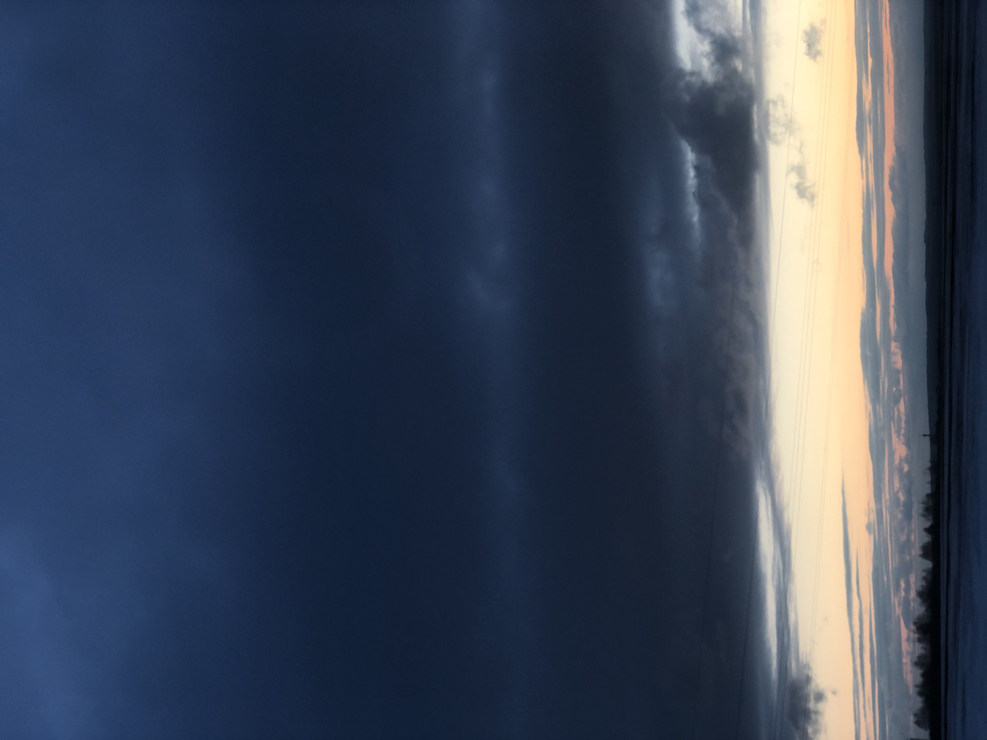 This landscape image, taken in Balakhta, Russia, presents a dramatic sky over what appears to be a large body of water and distant land. The scene is dominated by a stark contrast between dark, brooding clouds and a lighter, warmer horizon.
The left two-thirds of the image are enveloped in heavy, dark blue-grey clouds, which appear dense and potentially indicative of an overcast or stormy weather front. The texture of these clouds varies, with smoother, almost solid dark expanses blending into more textured, slightly lighter formations.
Towards the right side, the sky brightens significantly, transitioning into a band of pale yellow and soft orange hues near the horizon, suggesting either sunrise or sunset. Several thin, dark horizontal lines, resembling power lines or utility wires, are faintly visible stretching across this brighter section of the sky.
Below the horizon, on the far right, a dark, undulating line defines a distant treeline or landmass, appearing as a silhouetted stretch of nature. The area immediately beneath the warm-toned sky, extending to the bottom edge, seems to be a body of water, reflecting some of the ambient light and darkness.
The image captures a moment of powerful atmospheric conditions. There are no people, explicit activities, or visible text in the frame.