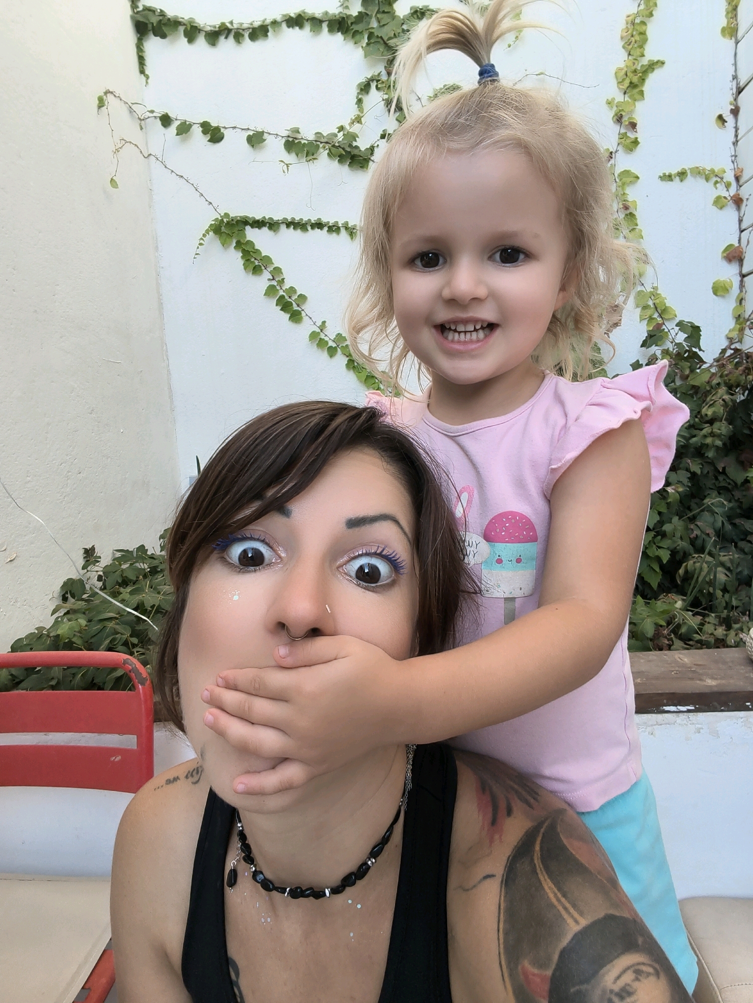 A young blonde girl with a ponytail stands behind a woman, covering the woman's mouth with her hand. The girl is wearing a pink t-shirt with an ice cream graphic and blue shorts. The woman has dark hair, blue eyeshadow, and a black necklace. She is wearing a black tank top and has tattoos on her arm and shoulder. They are outdoors, with a white wall and green ivy in the background. A red chair is visible to the left. The scene appears to be in a courtyard or patio area. The location is identified as Lachish Regional Council, Israel.