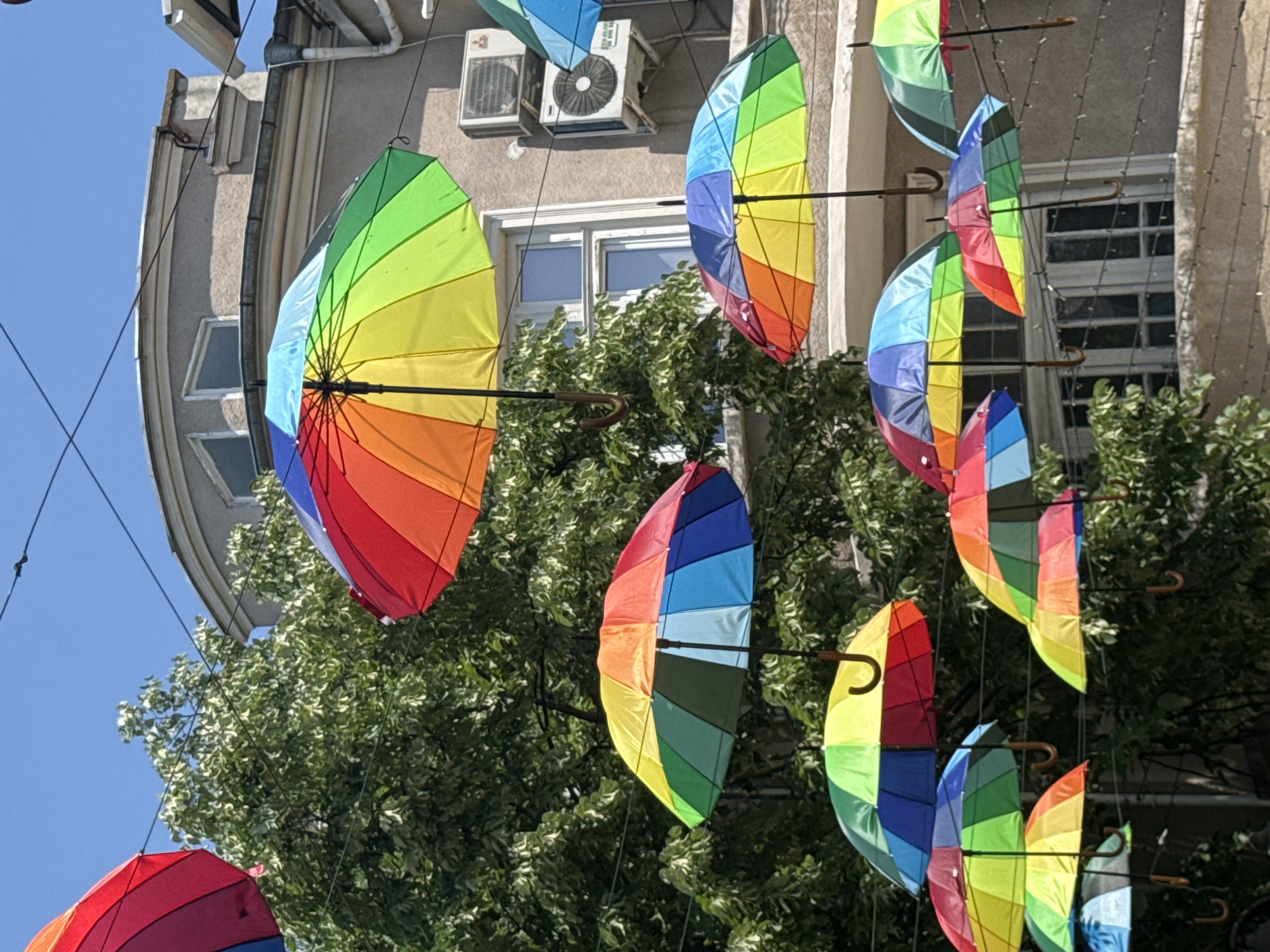 The image displays a vibrant street scene in Burgas, Bulgaria, characterized by an overhead display of numerous rainbow-colored umbrellas. These umbrellas, featuring segments of red, orange, yellow, green, blue, and violet, are partially open and suspended in multiple rows across the frame by thin black wires, creating a colorful canopy effect. Each umbrella has a visible curved handle.
In the background, a light-colored, multi-story building with a classic European architectural style is visible under a clear, bright blue sky. The building features several windows with white frames and at least two outdoor air conditioning units mounted on its facade. Lush green foliage from trees partially obscures parts of the background, adding natural elements to the urban setting.
The scene suggests a sunny daytime. The hanging umbrellas indicate a decorative installation, common in pedestrian zones to enhance the street's appeal. No discernible text is visible on any of the objects or surfaces.