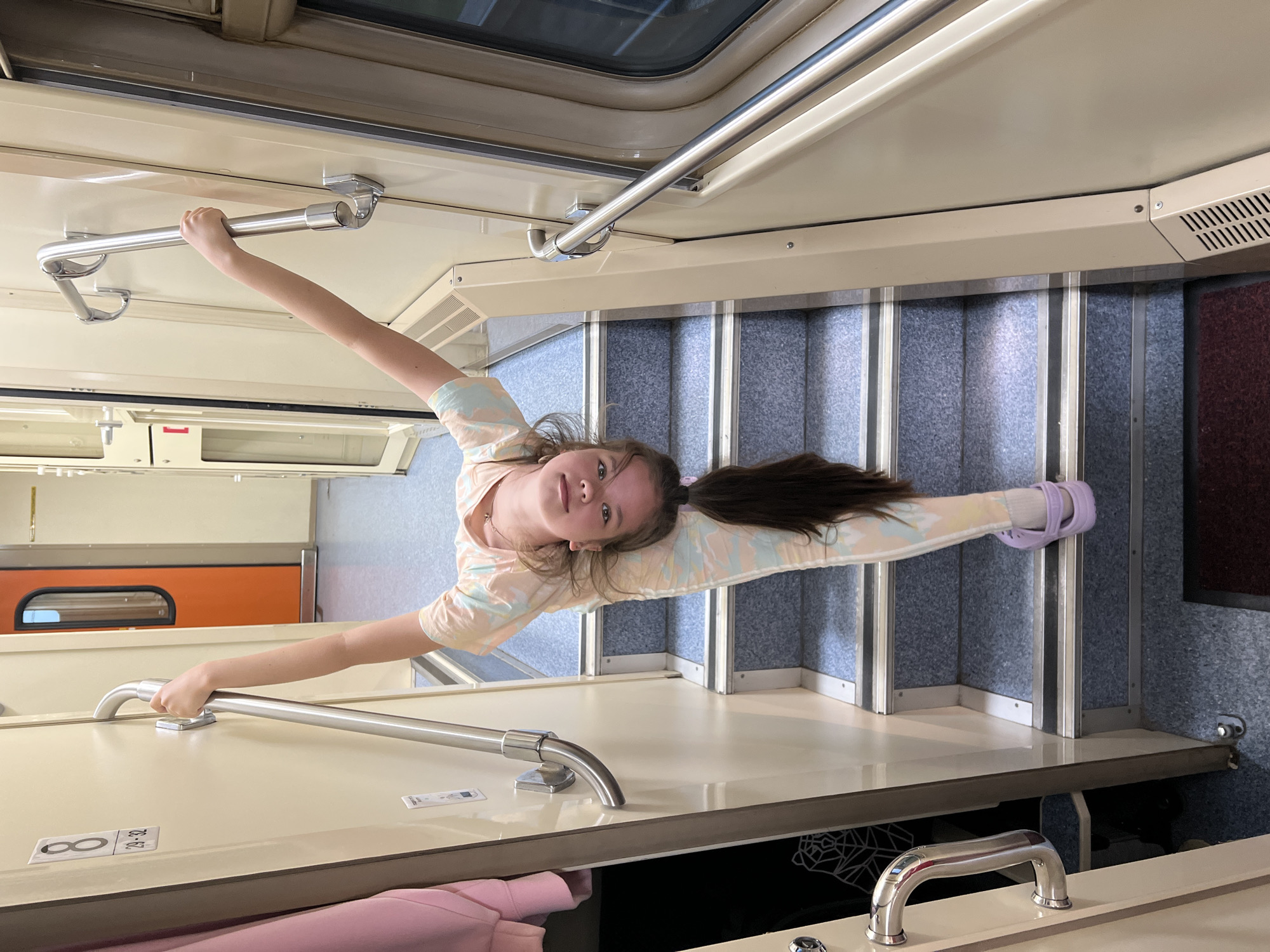 A young girl with long brown hair in a ponytail performs a split while holding onto handrails on the stairs of a train car. She is wearing a light-colored, patterned shirt and leggings, and light purple Crocs. The train car interior is cream-colored, with blue carpeted stairs and metal handrails. A closed orange door is visible in the background. The image does not provide any information or context to indicate that the location is Sarai-Chekurcha, Russia.