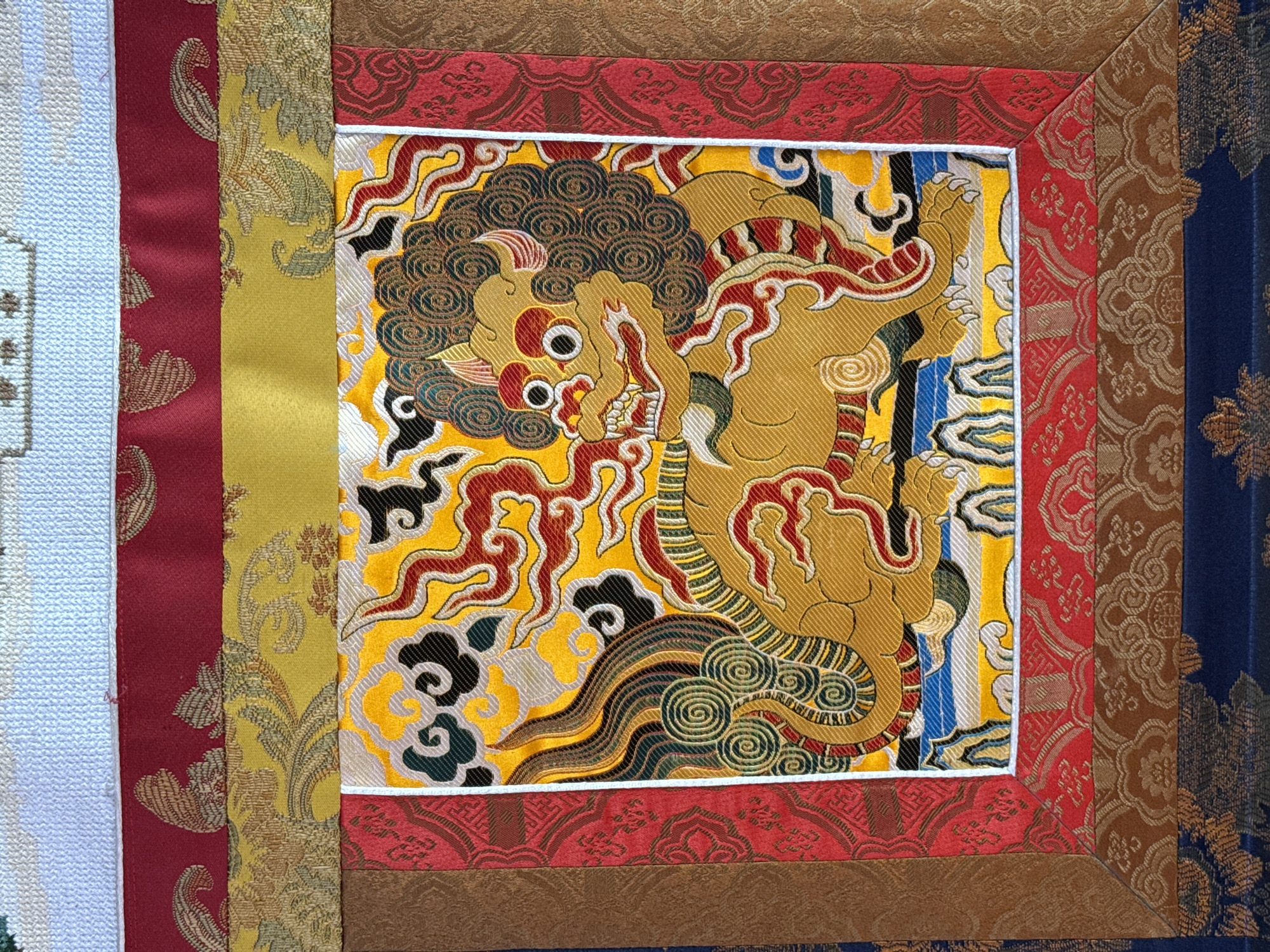 The image shows a square textile panel with a central depiction of a mythological creature, possibly a snow lion or similar guardian figure, rendered in gold, red, black, and green on a yellow background. The central panel is framed by a white border and then by a series of patterned fabric strips in red, brown, gold, and blue. The style and motifs suggest Tibetan or Himalayan art. There is no way to deduce a location or city from this single image.