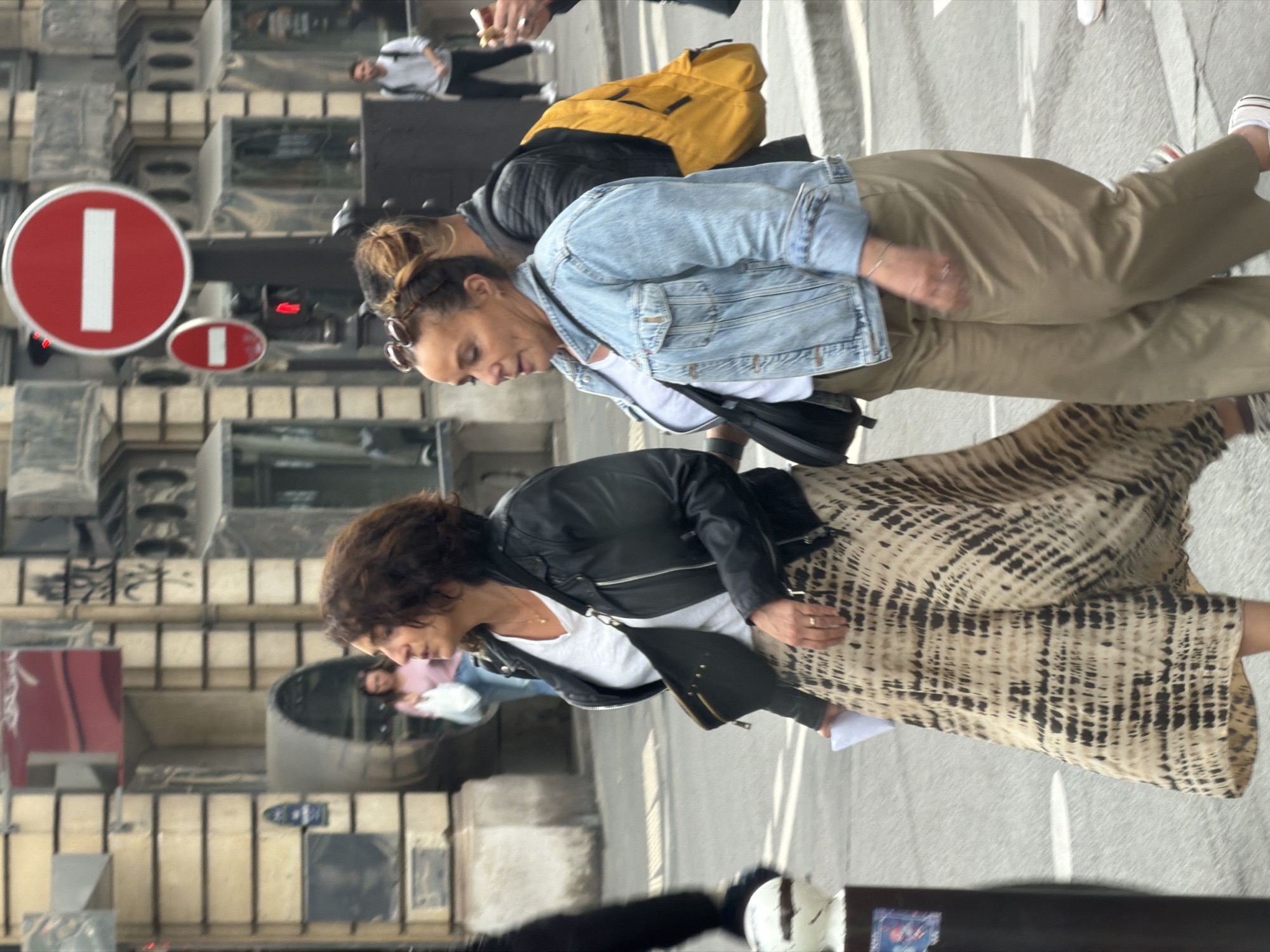 The image shows two women walking on a sidewalk in Paris, France. The woman in the foreground has short, curly dark hair and is wearing a black leather jacket over a white t-shirt, paired with a long beige and brown patterned skirt. She carries a black bag slung across her body. The second woman, slightly behind and to the right, has her hair tied back and is wearing a light blue denim jacket over a white top and khaki pants. She also has a black bag. A yellow backpack is visible on the back of another person partially visible. In the background, there is a street with a red and white "no entry" traffic sign. Buildings are visible in the background.