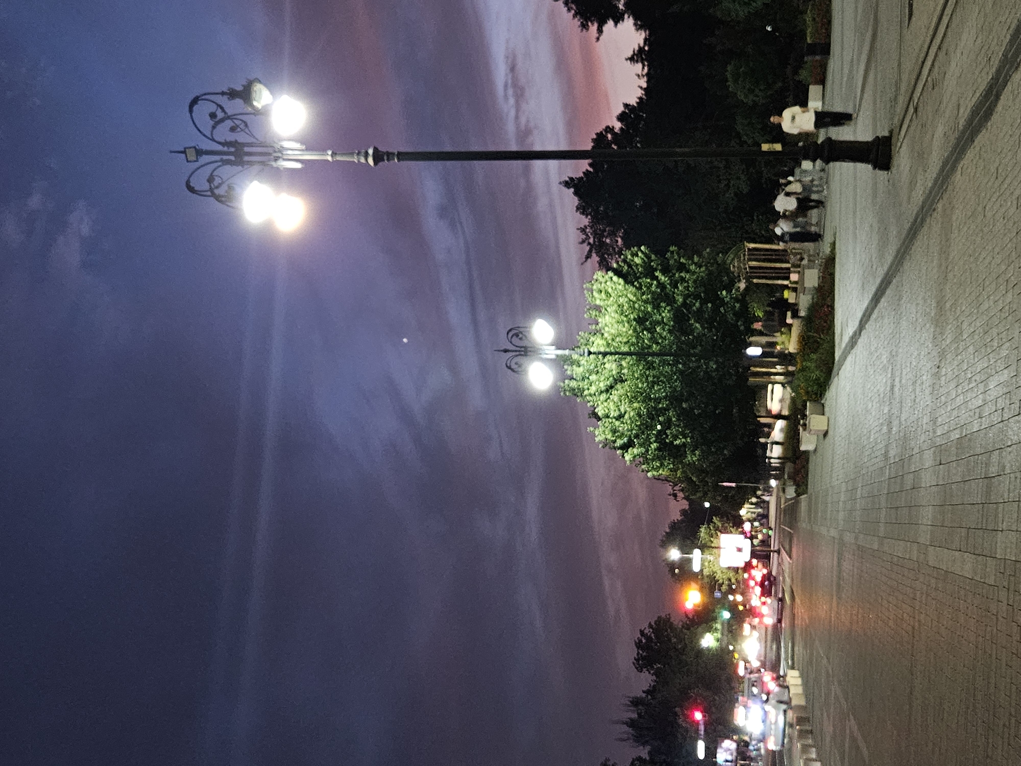 The image depicts a city street at dusk in Almaty, Kazakhstan, likely on an avenue such as Dostyk Avenue or Abay Avenue, given the characteristic street lighting and the presence of lush trees flanking the pedestrian walkway.
The sky is a deep twilight blue, with streaks of pink and purple from the setting sun visible near the horizon. The streetlights, in ornate traditional designs, are illuminated, casting a warm glow on the paved pedestrian walkway and the adjacent roadway. Several cars are on the road, their headlights and taillights creating streaks of light, suggesting movement.
On the pedestrian walkway, a few individuals are walking, some appearing to be strolling and others moving with purpose. One person in the foreground on the right is walking towards the viewer, wearing a white shirt and dark pants. To the left, near a gazebo-like structure and illuminated trees, a small group of people are gathered or walking. The trees are full and green, illuminated by the streetlights, providing a natural contrast to the urban environment. Concrete planters and bollards line the walkway, and some decorative fencing is visible. The overall impression is of a well-maintained urban space transitioning from day to night.