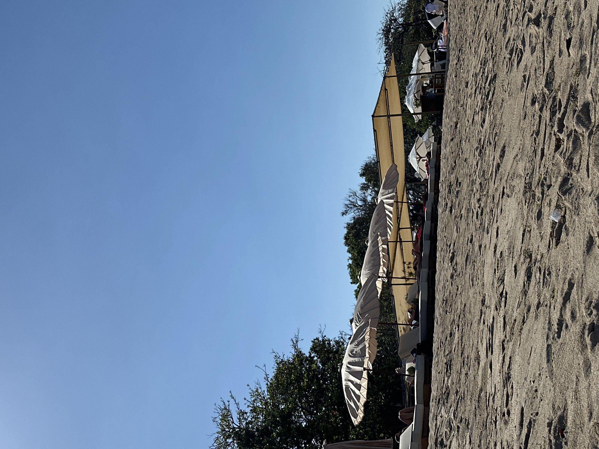 The image shows a beach scene in Kuta Selatan, Indonesia, on a clear, sunny day. The foreground is dominated by sand, with visible footprints and scattered debris. In the mid-ground, a row of lounge chairs and umbrellas are set up on the beach, with a few people reclining on the chairs. The umbrellas are a mix of striped and solid colors, providing shade. Behind the lounge chairs, there are trees and some structures, including what appears to be a bar area with more umbrellas and seating. In the far right of the image, a group of people are gathered, some of whom are dressed in white. The background consists of a clear blue sky.