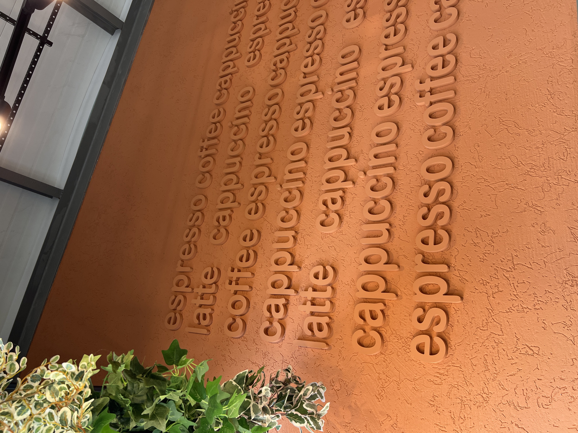 A close-up, angled shot shows an orange textured wall with raised, light orange letters that spell out coffee-related terms. The words appear in rows, repeating "espresso," "latte," "coffee," and "cappuccino." At the top left, a section of green ivy with variegated leaves hangs into the frame, partially obscuring the upper portion of the wall and letters. The background consists of a white ceiling with dark gray structural beams and lights. The lighting is bright, casting subtle shadows from the raised letters. The scene is an interior, likely a cafe or coffee shop, devoid of any people or explicit actions.