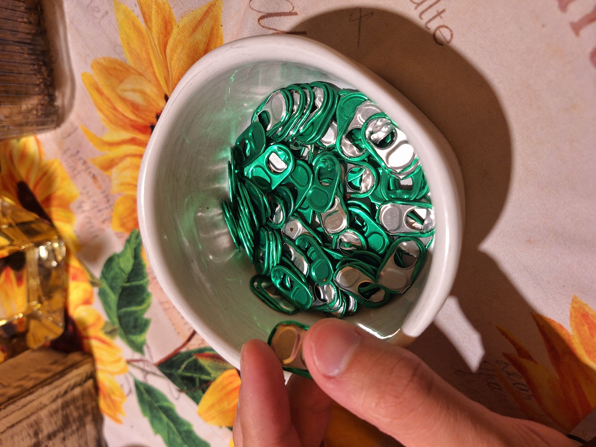 A high-angle, indoor shot displays a collection of green and silver soda can tabs filling a white, asymmetrical bowl. A person's hand is visible in the lower right corner, holding a single tab. The bowl sits on a patterned tablecloth with floral designs, including sunflowers. The tablecloth covers a wooden surface, partially visible on the left side of the frame. The environment appears to be someone's home. The image does not contain any information to determine a location in Sofia, Bulgaria.