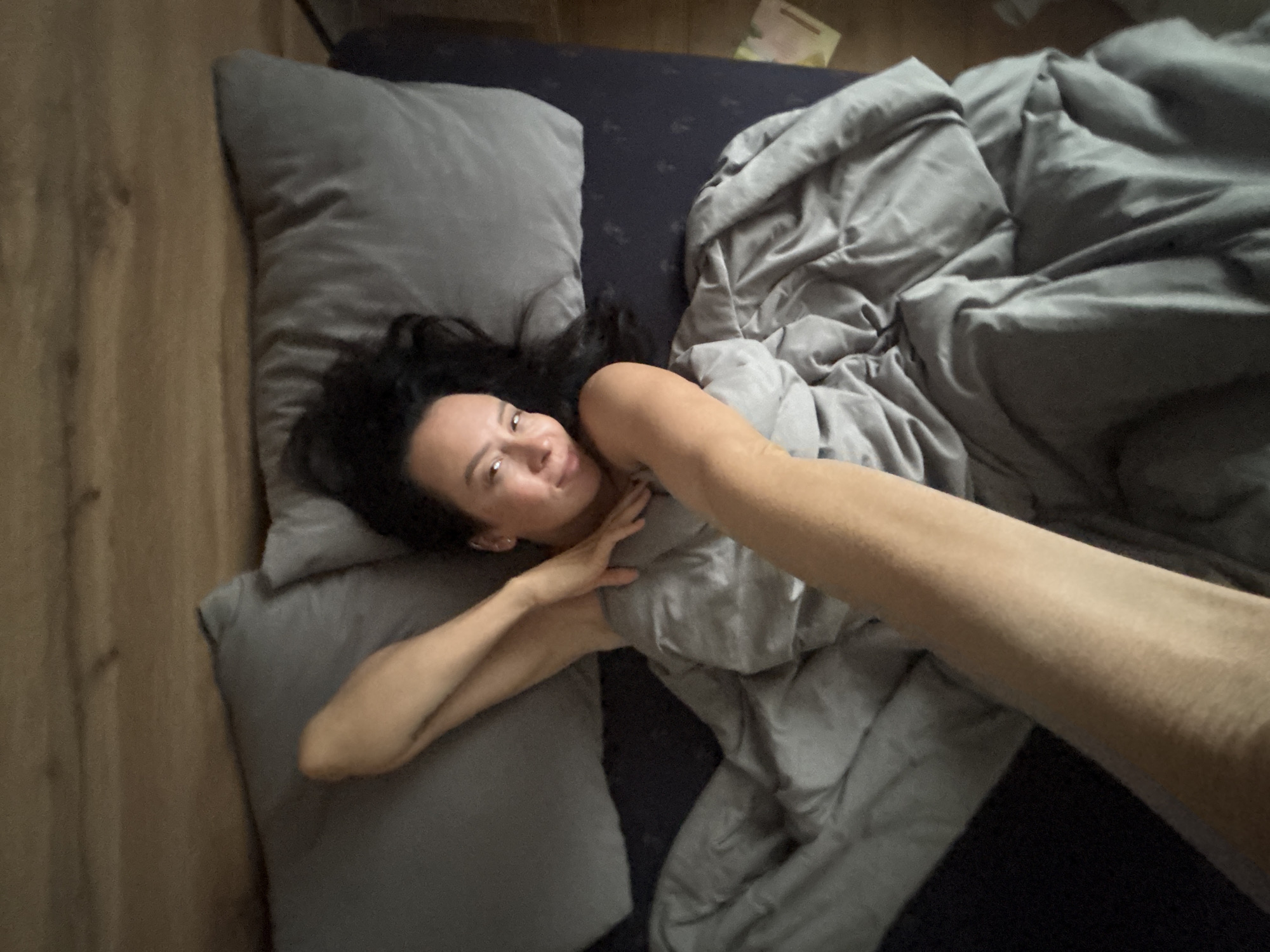 A woman with dark hair is lying on a gray bed with gray pillows and a gray comforter. Her left arm is extended towards the camera. She is looking towards the camera. The photo appears to be taken in a room with wooden flooring. There is no evidence in the image to suggest the location is Chaikovsky, Russia.