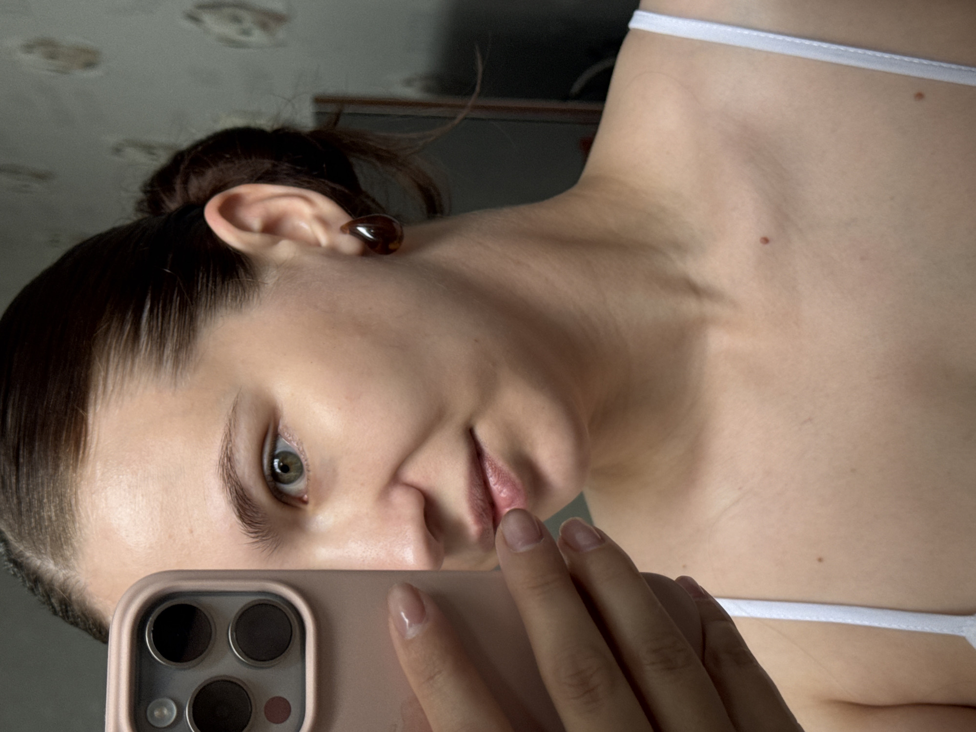 A young woman with her hair pulled back in a bun is taking a selfie with a smartphone in her left hand. She is wearing a white tank top. The woman has light skin and green eyes. She is wearing dark brown teardrop-shaped earrings. In the background, a patterned wallpaper is visible. The image appears to have been taken indoors. There is no information about the specific location within the city of Chaikovsky, Russia.