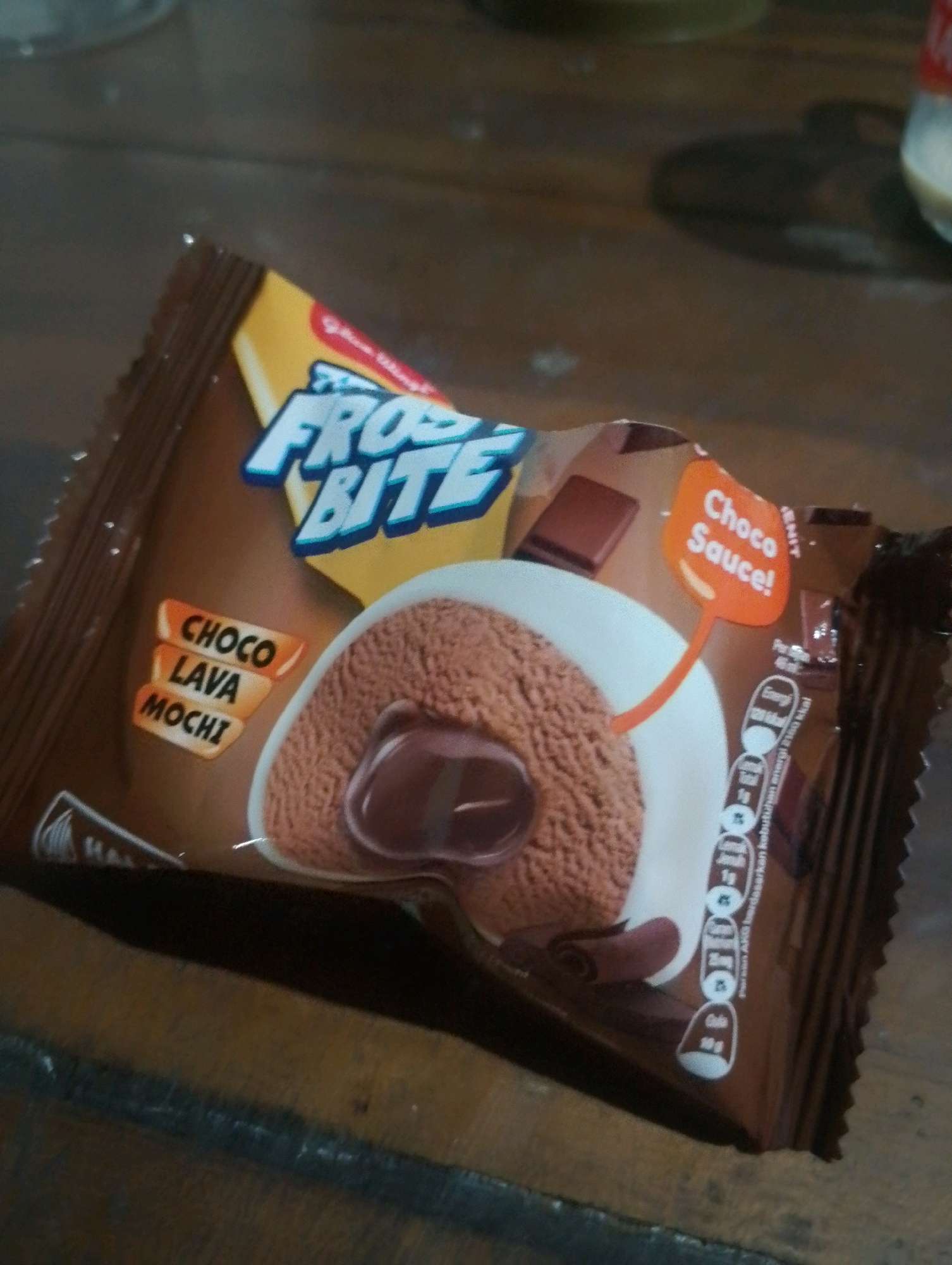 A chocolate-flavored mochi dessert, branded "Frost Bite," is presented on a wooden surface, likely a table. The packaging is predominantly brown, featuring prominent "Frost Bite" lettering in a stylized font, and a depiction of the mochi itself with chocolate sauce. Text on the packaging indicates "Choco Lava Mochi" and "Choco Sauce!". The lighting suggests an indoor setting, possibly a home or a small shop, with diffused light casting a shadow on the wooden surface. The overall impression is of a product shot or a casual photograph taken in a residential environment. This scene could be located in Kendalrejo, Indonesia, given the context.