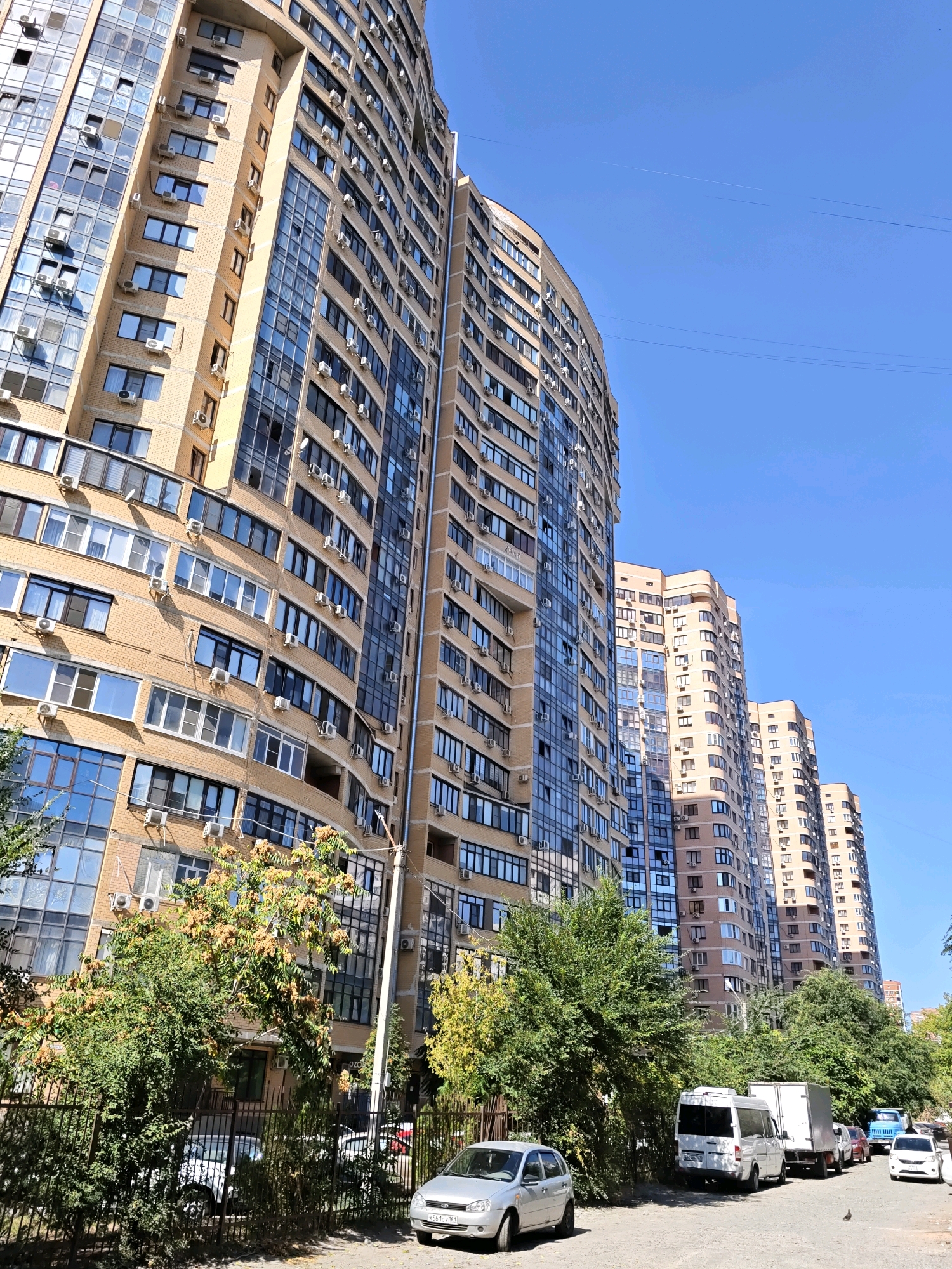 The image depicts a street scene in Rostov-on-Don, Russia, characterized by towering, modern apartment buildings with light brown brick facades and numerous windows. The buildings are curvilinear in design, creating a sense of depth and scale. Air conditioning units are visible attached to many of the windows. In the foreground, a street lined with trees and a black metal fence separates the sidewalk from the buildings. Several vehicles are parked along the street, including a silver sedan, a white minivan, a white truck, and a blue truck. The sky is clear and blue, indicating a sunny day.