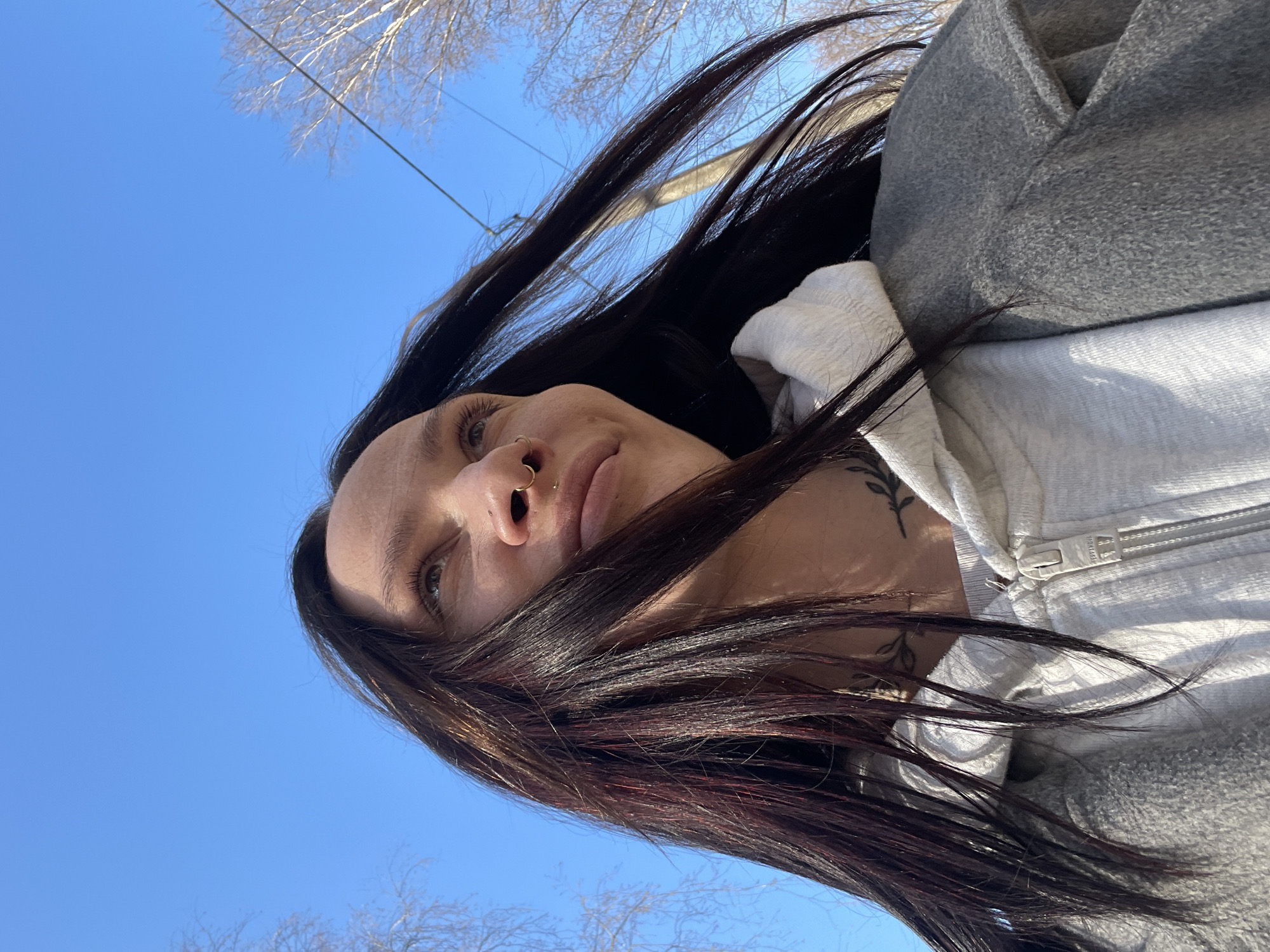 A woman with long, dark hair, a nose ring, and a tattoo on her neck is seen from a low angle against a clear blue sky. She is wearing a white hooded sweatshirt and a gray jacket. Bare trees are visible in the background. There is no verifiable evidence to suggest this image was taken in Izhevsk, Russia.