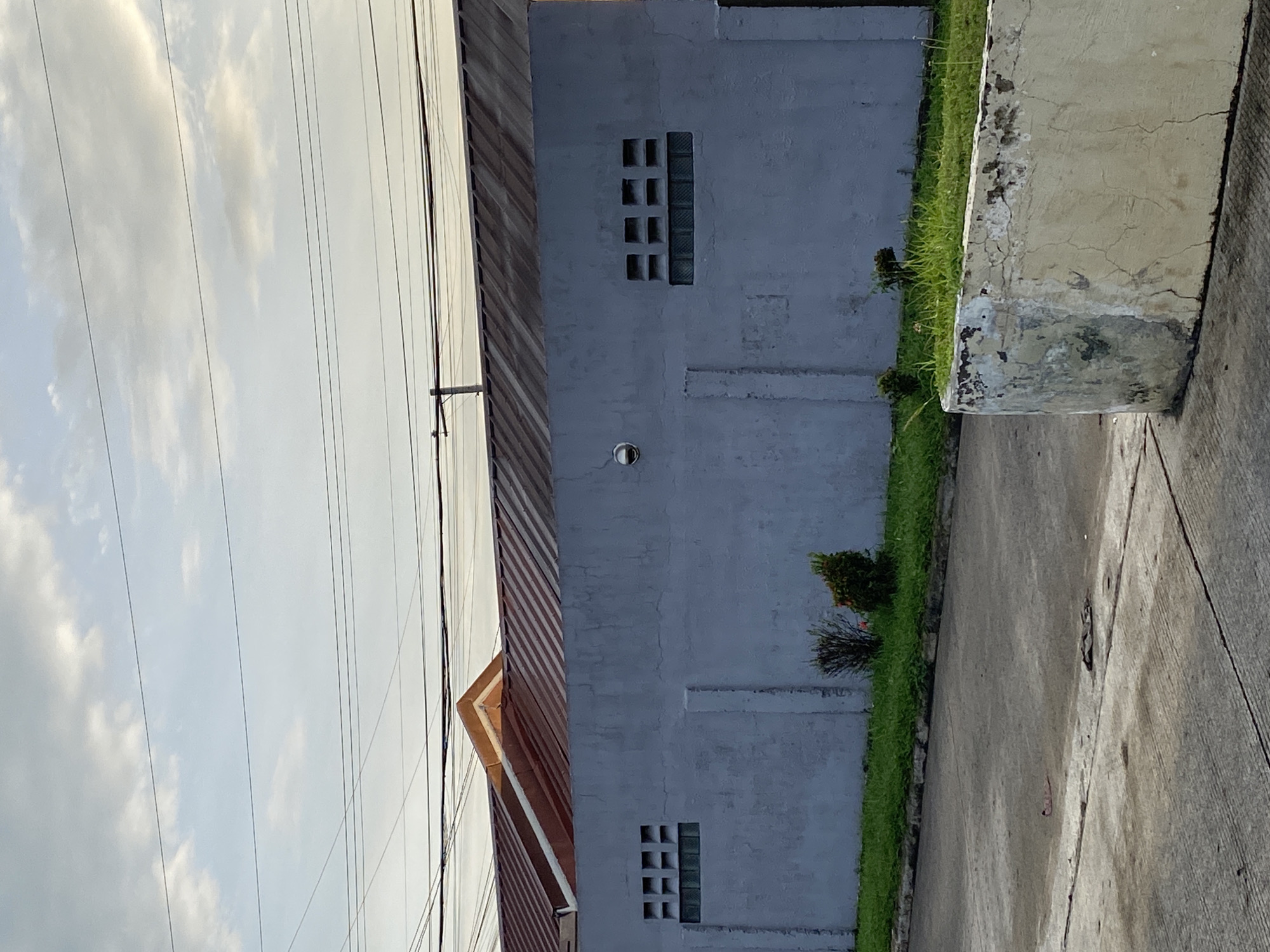 The image, captured in Davao City, Philippines, depicts an outdoor scene featuring a building, ground, and sky. The central subject is a pale blue-grey concrete building with a prominent brown and reddish-orange corrugated metal roof, dominating the left side of the frame. The building's wall features two distinct sets of rectangular openings: one near the top and another near the bottom. Each set includes several small square vents and a vertical section of glass blocks, suggesting ventilation or decorative elements. A small, dark fixture is mounted on the wall between these openings.
In the foreground, a textured, light-colored concrete block or planter, exhibiting cracks and stains, runs along the right side. Below the building and planter, a vibrant strip of green grass grows, punctuated by a couple of small, bushy plants. A light grey concrete pathway or road occupies the bottom right portion of the frame. Above the building, a bright, cloudy sky is visible, crisscrossed by numerous thin, dark utility wires, likely power lines.
The scene is static, with no human activity or specific event taking place. The soft, diffused lighting suggests it is daytime, possibly early morning or late afternoon, under an overcast sky. The simple concrete architecture and corrugated roof are common in the region. There is no visible text.