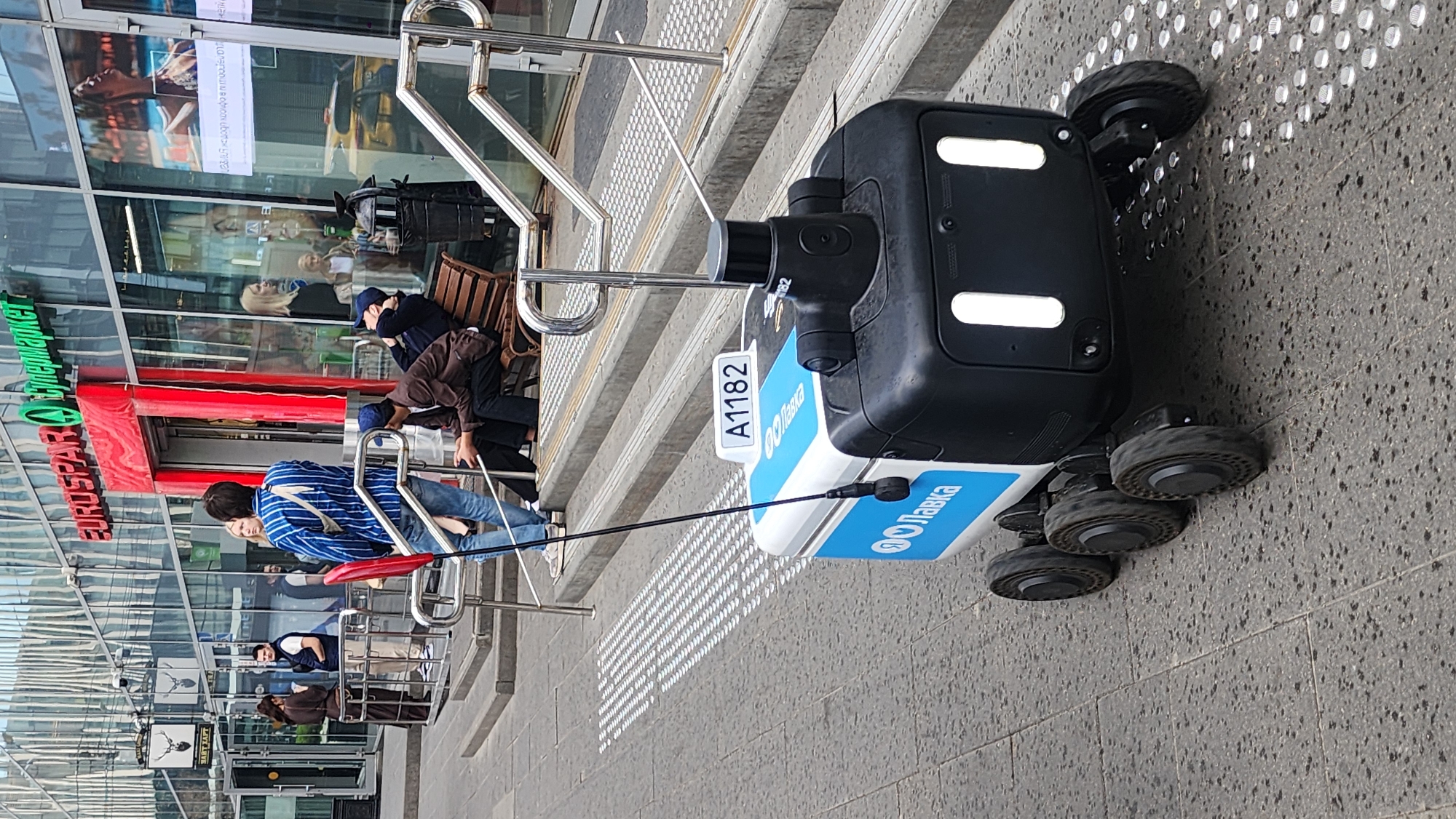 The image depicts a delivery robot standing on a sidewalk next to a building in what appears to be Moscow, Russia. The robot is branded with "Яндекс Лавка" (Yandex Lavka), a grocery delivery service. It has a black upper section with white lights and a blue and white lower section. The robot is near a set of stairs leading to a building entrance. People are visible in the background, some sitting on a bench. The building has the sign "EUROSPAR Ситимаркет" (EUROSPAR Citymarket). The sidewalk has tactile paving for the visually impaired.
