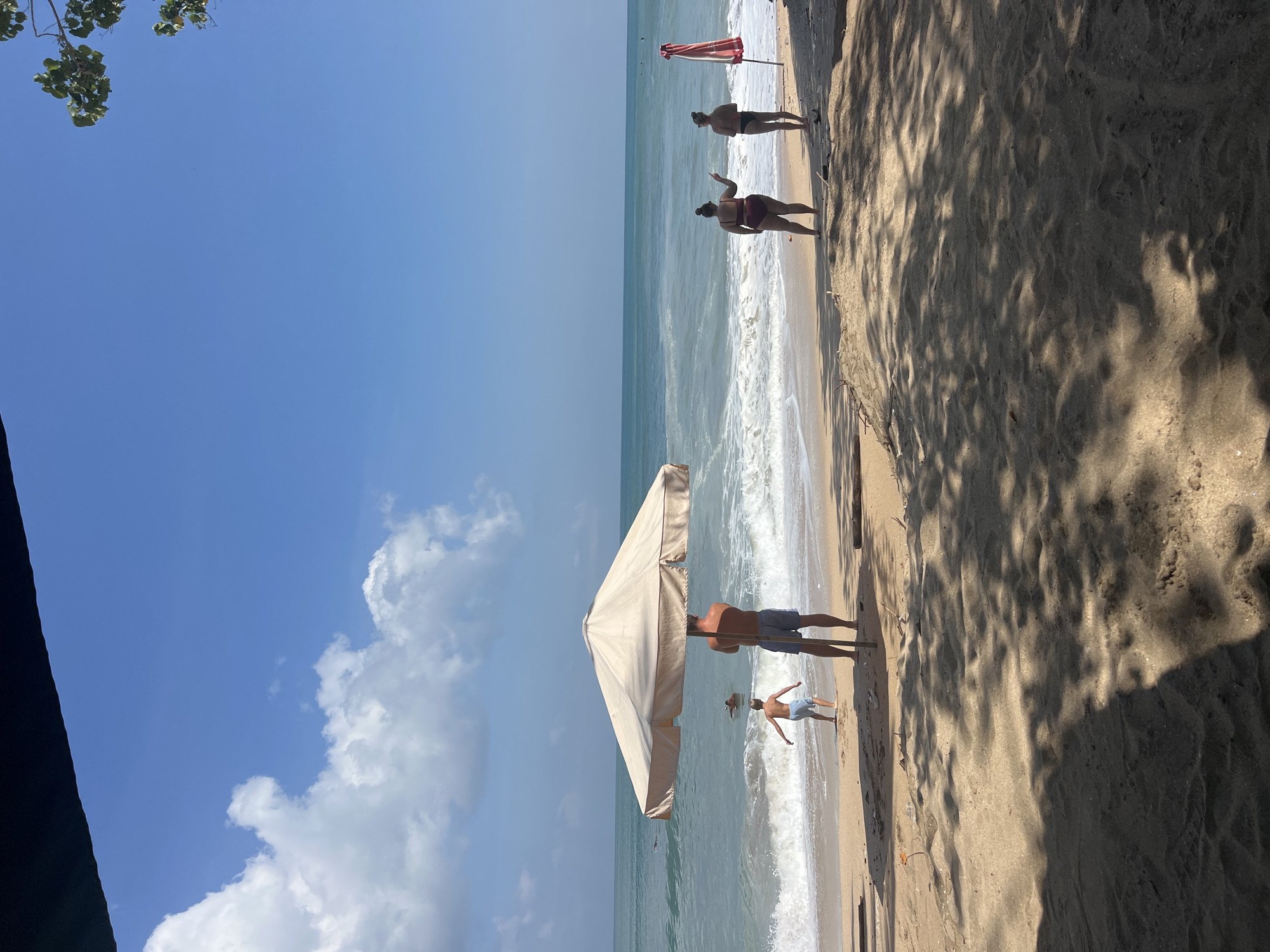 This is a horizontal, eye-level, medium shot of a beach in Kuta, Indonesia, on a sunny day. The ocean is calm with small waves breaking on the shore. The sky is blue with a few white clouds. In the foreground, a sandy beach is dotted with shadows of palm trees. A large, tan umbrella is set up on the sand. A man in blue shorts stands under the umbrella, facing away from the camera. To his left, a young boy in blue shorts runs towards the water. Further down the beach, two women stand near a red and white striped umbrella. The woman on the left, wearing a dark red swimsuit, points towards the ocean. The woman on the right, wearing a black bikini, stands beside her. In the distance, a few surfers can be seen in the water.