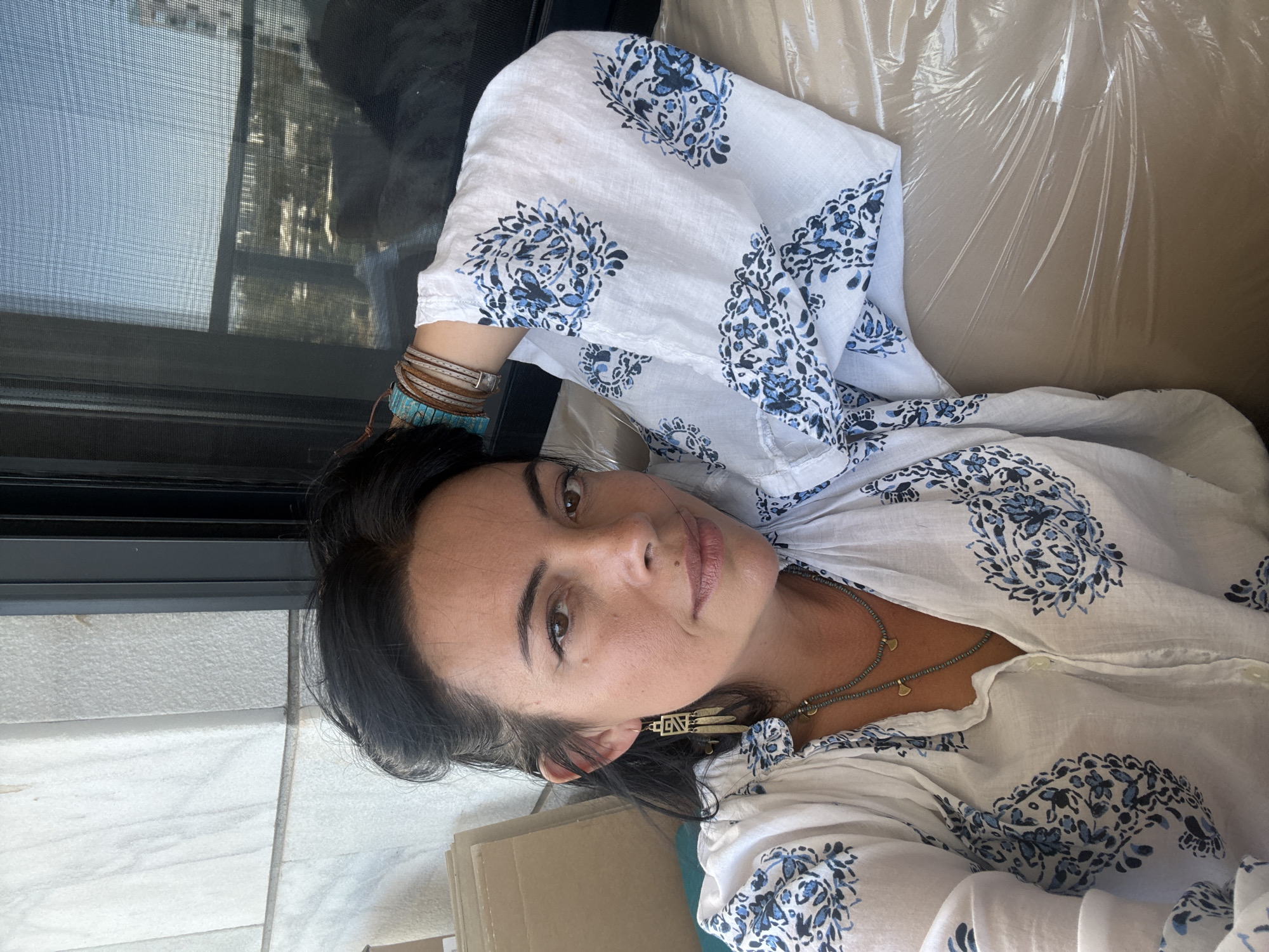 A woman with dark hair and brown eyes is photographed from the chest up, lying down and looking at the camera. She is wearing a white blouse with blue floral patterns and two layered necklaces. She has earrings and bracelets on her left arm, which is resting behind her head. She appears to be indoors, possibly in an apartment in Or Yehuda, Israel, with a window showing a cityscape in the background. The room is sparsely furnished, with a beige sofa and some cardboard boxes visible.
