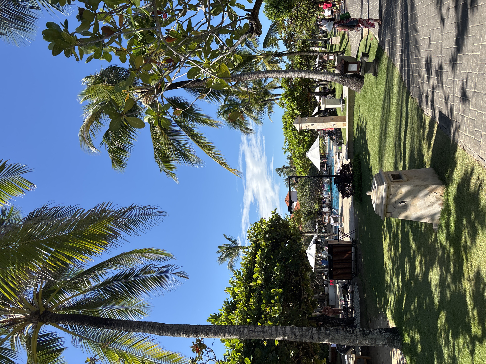 The image shows a tropical scene in Nusa Dua, Indonesia. In the background, there is a resort area with a swimming pool, lounge chairs, and white sun umbrellas. Lush green vegetation, including palm trees and other trees, is prominent, framing the view. A stone pathway runs along the right side, bordering a green lawn. There are people walking on the pathway in the distance. The sky is blue with some wispy clouds. The overall impression is a sunny and relaxed tropical atmosphere.