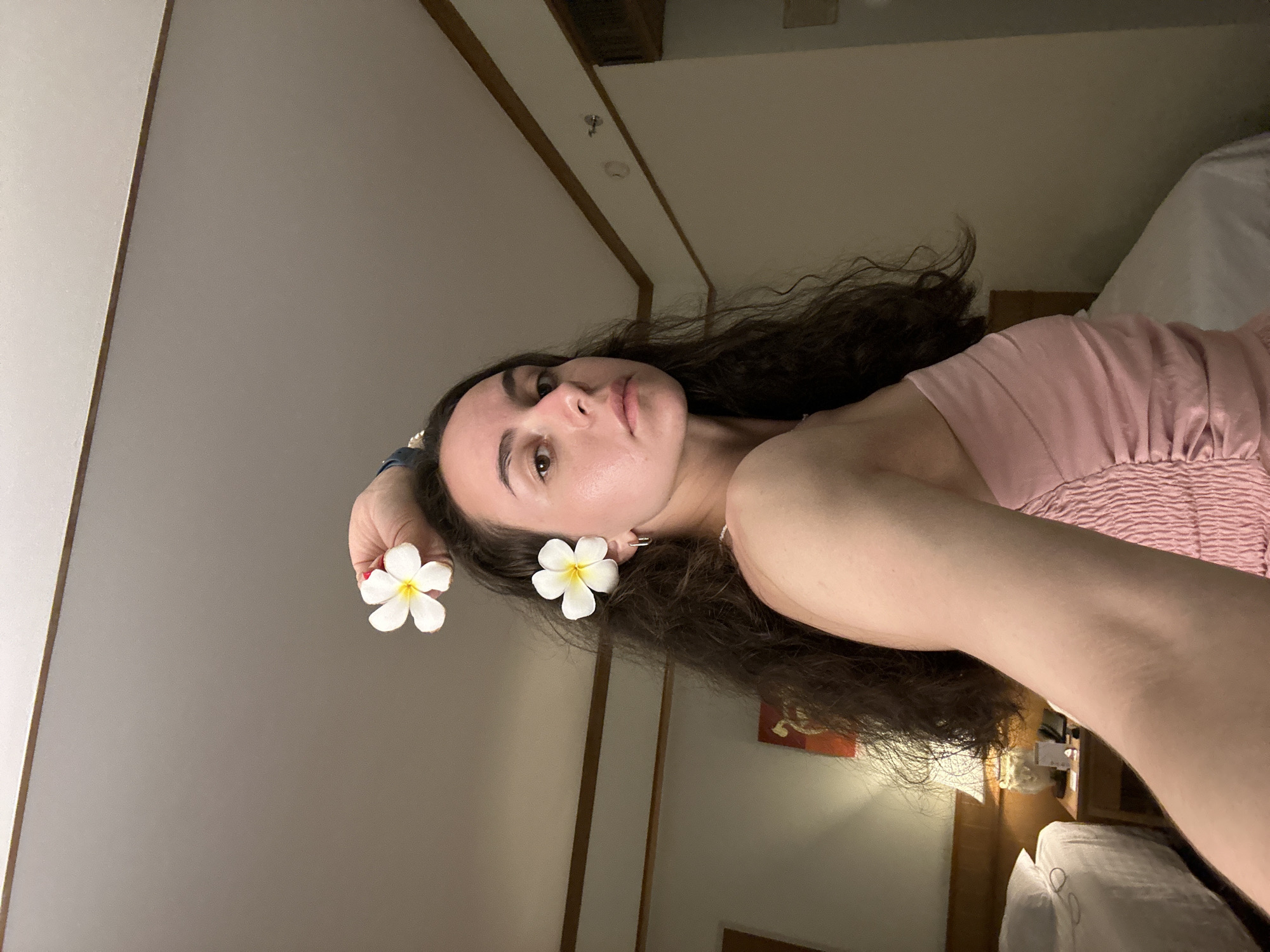 A person is lying on a bed in a room, likely in Pattaya City, Thailand. The person has long, dark, curly hair and is wearing a pink top. They are holding two white frangipani flowers, one near their ear and the other near their hand. The room has a ceiling with wooden accents and lighting fixtures. A bed with white linens is visible in the background, along with a bedside table.