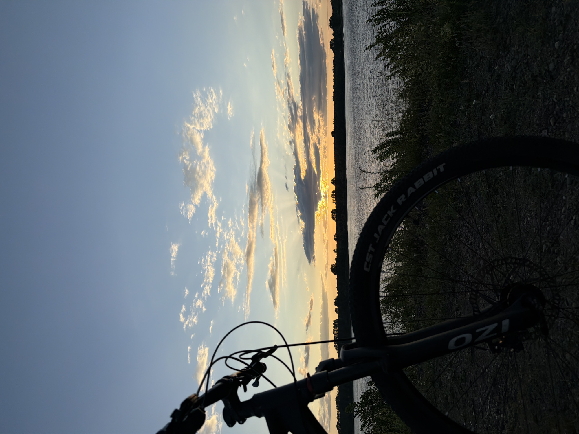 This is a medium shot, landscape orientation, photograph taken at eye level of a bicycle in the foreground, parked on a dirt path. The bicycle is facing right and partially obscures the view of a calm body of water in the mid-ground. The water is bordered by a dark, tree-lined horizon under a vast sky at sunset. The sky is filled with scattered clouds, tinged with orange and yellow hues near the horizon, indicating the sun is setting. Sunlight rays are visible piercing through some of the clouds. The overall atmosphere is serene. The location is specified as Chaikovsky, Russia, a city situated near the Kama River.