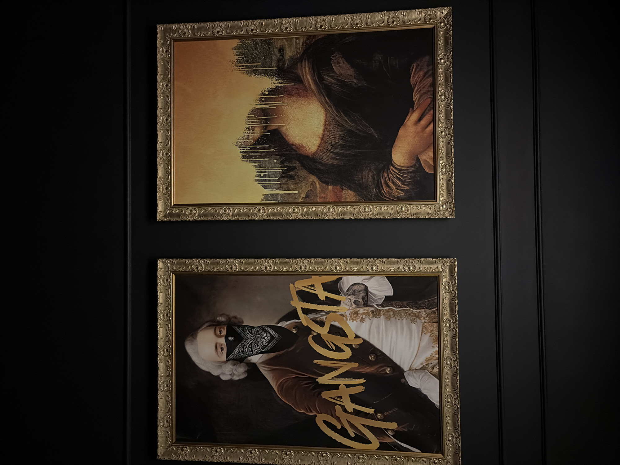 The image shows two framed artworks hanging on a dark, possibly black, vertically paneled wall. The top artwork depicts a deconstructed Mona Lisa with a dripping effect covering part of her face. The bottom artwork is of a classically dressed person wearing a bandana over their face and the word "GANGSTA" written diagonally across the image in gold lettering. The city of Chaikovsky, Russia, is not evident in the image.