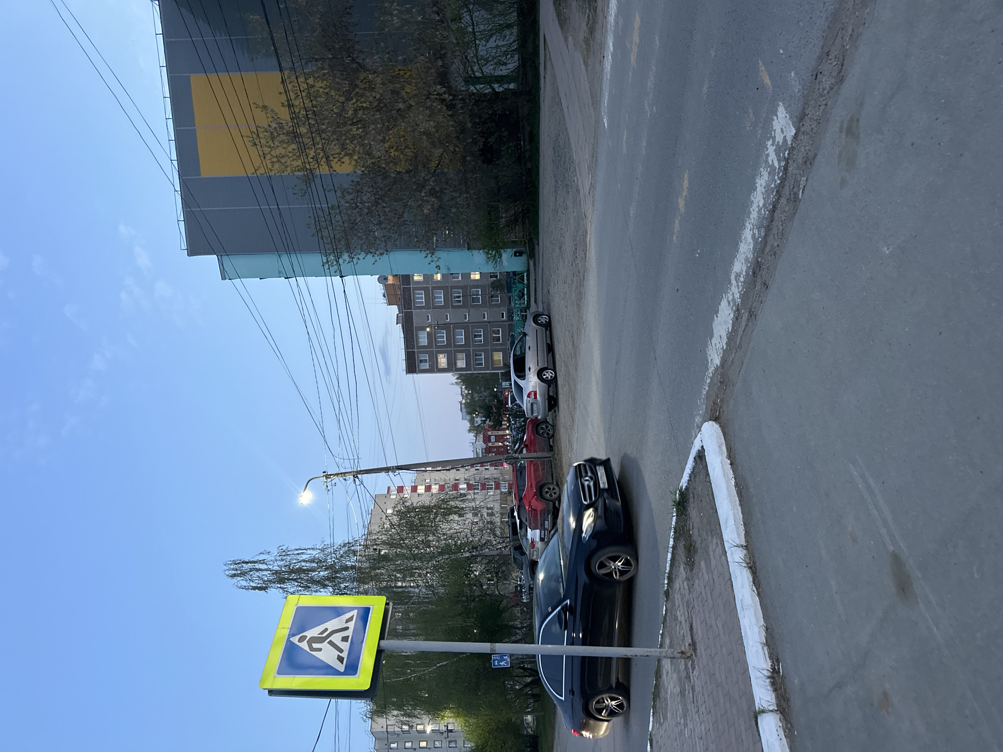 The image captures an urban scene in Chaikovsky, Russia, likely taken in the early evening. A pedestrian crossing sign is prominently displayed on the left side of the frame, indicating a designated crosswalk. A line of parked cars occupies the right side of the road. Buildings of varying heights and architectural styles line the street, reflecting the city's urban landscape. Overhead power lines crisscross the sky. The sky is partially visible, suggesting the image was taken during daytime or twilight hours. The overall atmosphere appears calm and ordinary, capturing a typical street view.