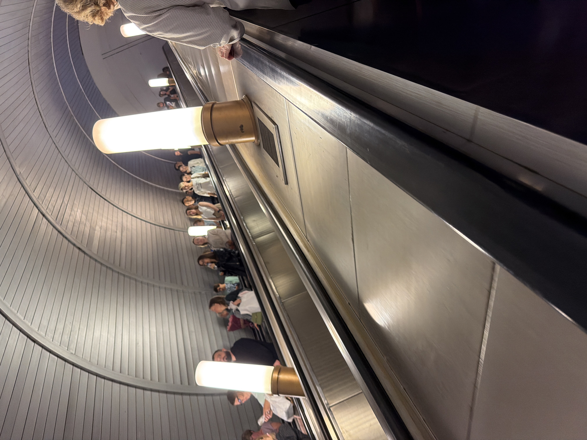 The image shows a view looking down an escalator in a Moscow Metro station. Several people are visible on the escalator, facing upwards. The escalator is constructed of shiny metal and is illuminated by tall, cylindrical light fixtures. The ceiling of the station is curved and lined with horizontal elements, typical of some Moscow Metro stations. The overall scene is one of transit and public transportation in an urban environment.