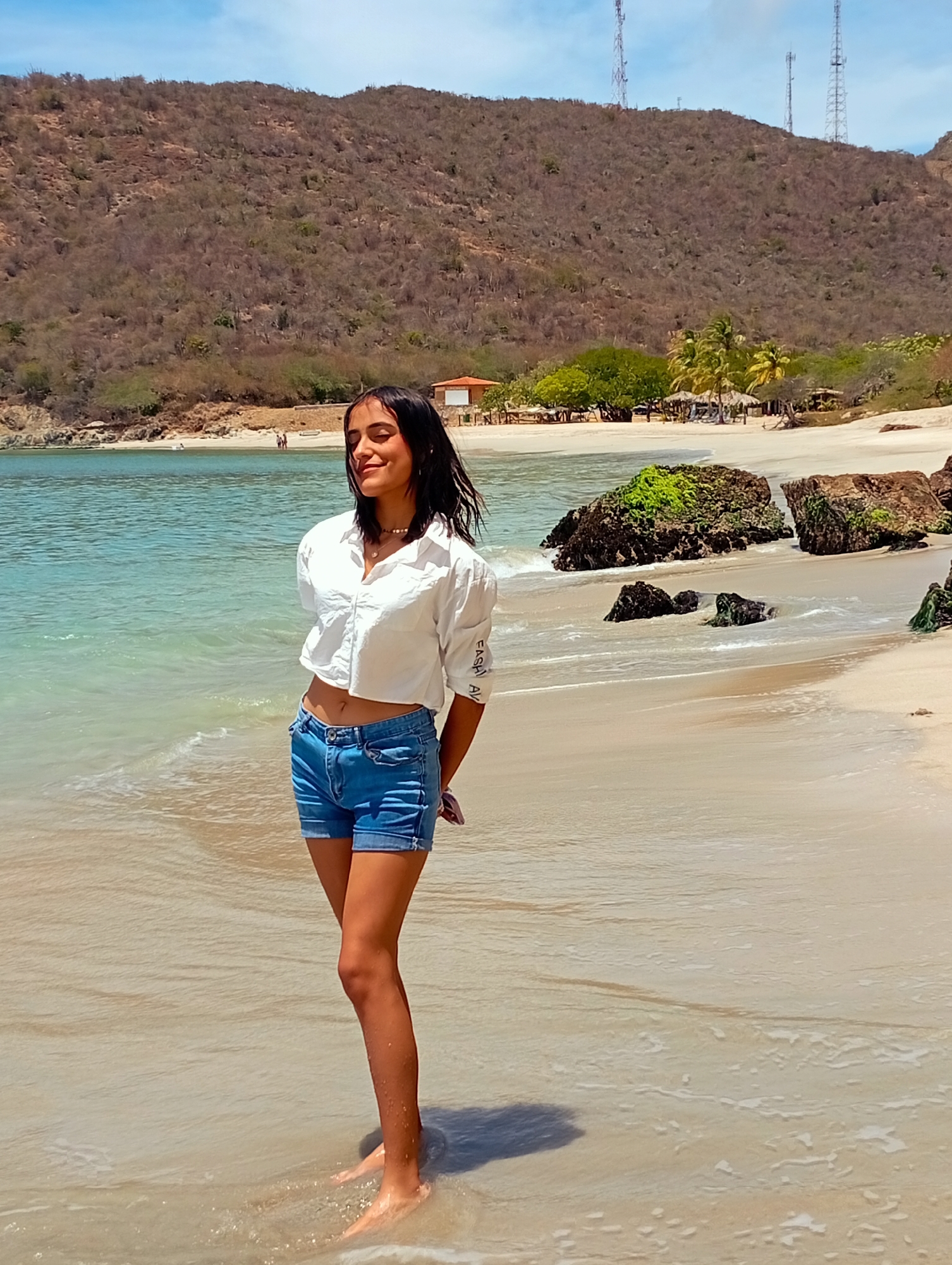 The image displays a young woman standing on a sandy beach in Pedro González, Venezuela, under a bright, sunny sky. She is positioned centrally, facing slightly away from the camera, with her eyes closed and a serene smile, basking in the sun. She has dark hair and is wearing a white cropped long-sleeve shirt and light blue denim shorts, with her bare feet immersed in the clear, shallow turquoise water at the edge of the shore. Her hands are clasped behind her back.
The scene features light-colored sand and several dark, moss-covered rocks in the foreground and midground, both in and out of the water. The calm ocean stretches into the distance. In the background, a large, arid mountain covered in dry, sparse vegetation rises, but closer to the beach, there are patches of lush green trees, including palm trees, and several small, rustic buildings or beach huts. Three tall cell phone towers are visible on the distant mountain ridge against a blue sky with light clouds. The visible text "FASHION AVE" can be faintly discerned on the cuff of her right sleeve. The overall atmosphere is one of relaxation and natural beauty on a clear, warm day.