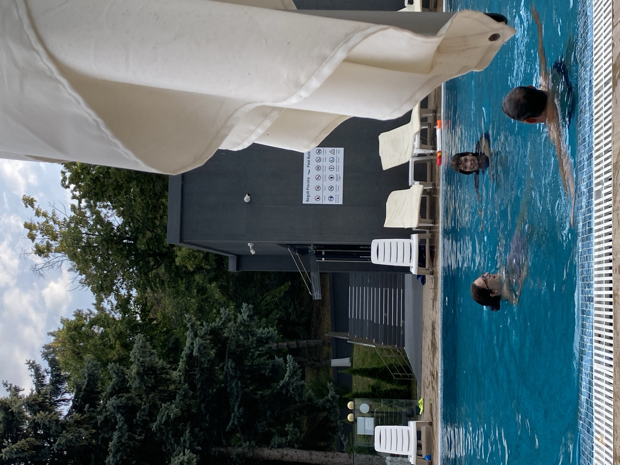 In Ustia, Moldova, a group of people are enjoying a swim in a bright blue outdoor pool. The water is clear and inviting, reflecting the overcast sky above. In the foreground, a woman with glasses and dark hair smiles as she paddles through the water. Behind her, a young boy with a broad smile is also swimming. Further back, a man with a bald head and dark hair is swimming with his arms outstretched. The pool area is surrounded by lush green trees, creating a serene and natural ambiance. White lounge chairs are neatly arranged around the pool, suggesting a place for relaxation. A modern, dark-colored building is visible in the background, with a sign displaying "Reguli Piscina Pool Rules" with various pictograms. The overall scene captures a moment of leisure and enjoyment in a tranquil outdoor setting.