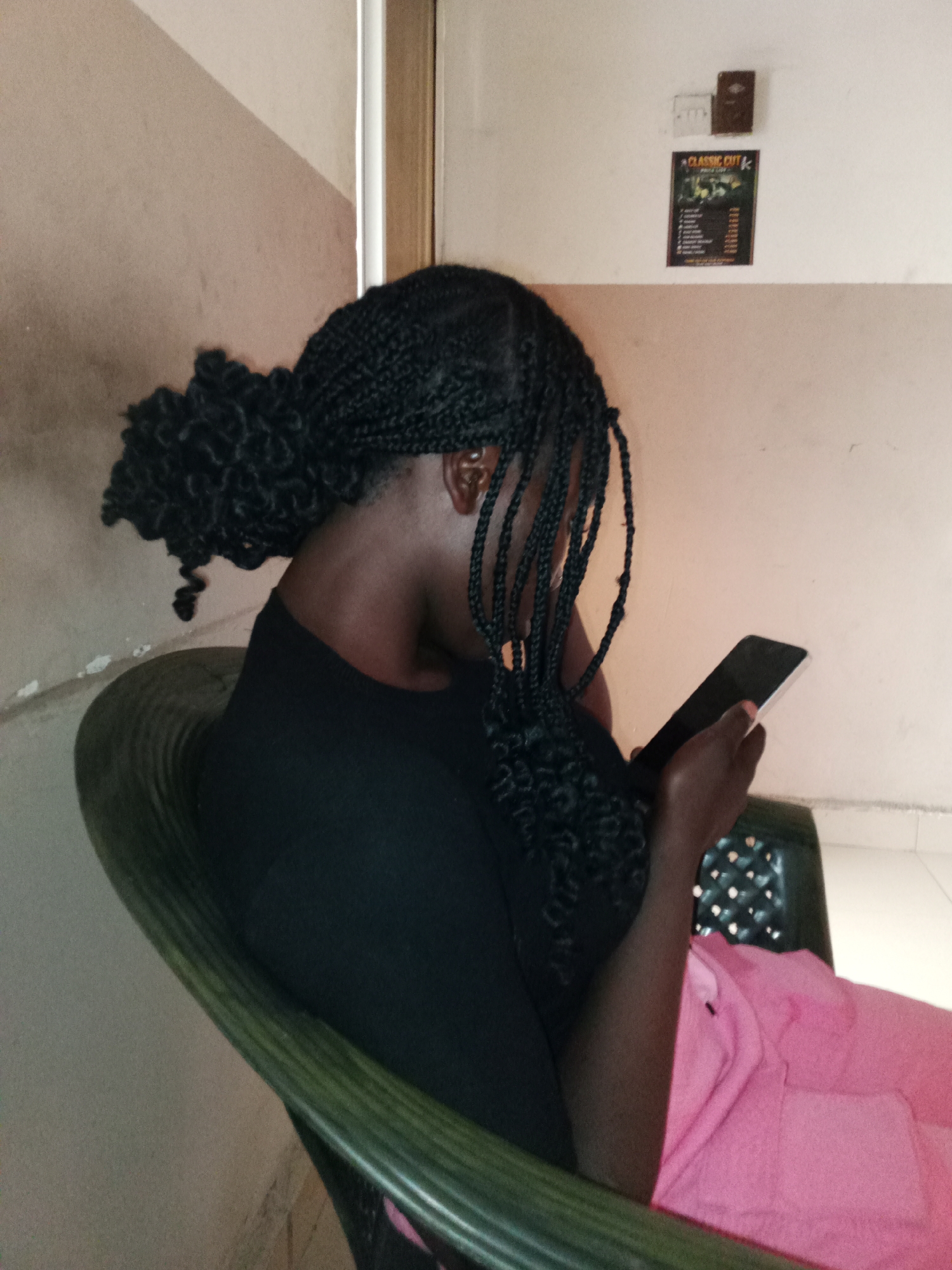 A young Black female is seated in a green plastic chair, facing away from the camera and looking at her smartphone. Her braided hair is pulled back into a bun with loose braids framing her face and shoulders. She is wearing a black t-shirt and a pink garment over her lap. The environment appears to be indoors, with a textured beige wall behind her and a white vertical pillar to her right. A framed poster with text and images, along with two small electrical outlets, are visible on the wall. The lighting suggests an indoor setting. The image is cropped to focus on the individual and her activity. The scene is likely located in Jalingo, Nigeria, based on the request.