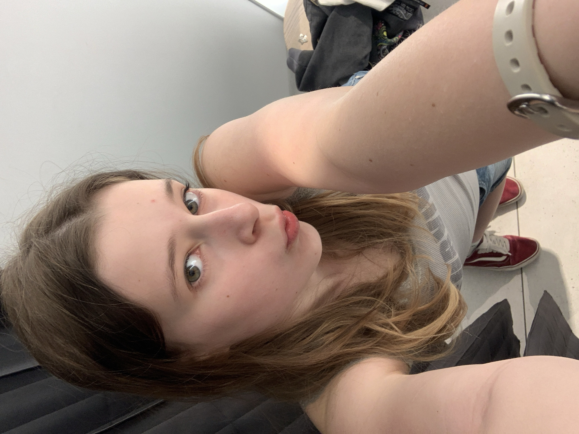 A medium shot, taken as a selfie, captures a young person indoors, likely in Belgrade, Serbia. The subject, a female with light brown hair, fair skin, and striking green eyes, looks directly at the camera with a slightly wide-eyed, curious expression and softly pursed lips. Her hair cascades over her shoulders, framing her face.
She is wearing a grey and white striped tank top or t-shirt, paired with blue denim shorts. A white fitness tracker or watch with a silver buckle is visible on her right wrist. Her left arm, extended to hold the camera, is partially seen in the upper right. Her footwear consists of red canvas sneakers with white laces, visible on the light-colored tiled floor.
The setting appears to be a simple, well-lit room, possibly a changing room or a retail space, given the plain light grey walls. In the background, behind her, a pile of dark clothing or fabric is visible, along with a darker, possibly pleated, element near the bottom left corner. The lighting is bright and even, indicating an indoor environment, but the time of day is not discernible. There is no visible text in the image.