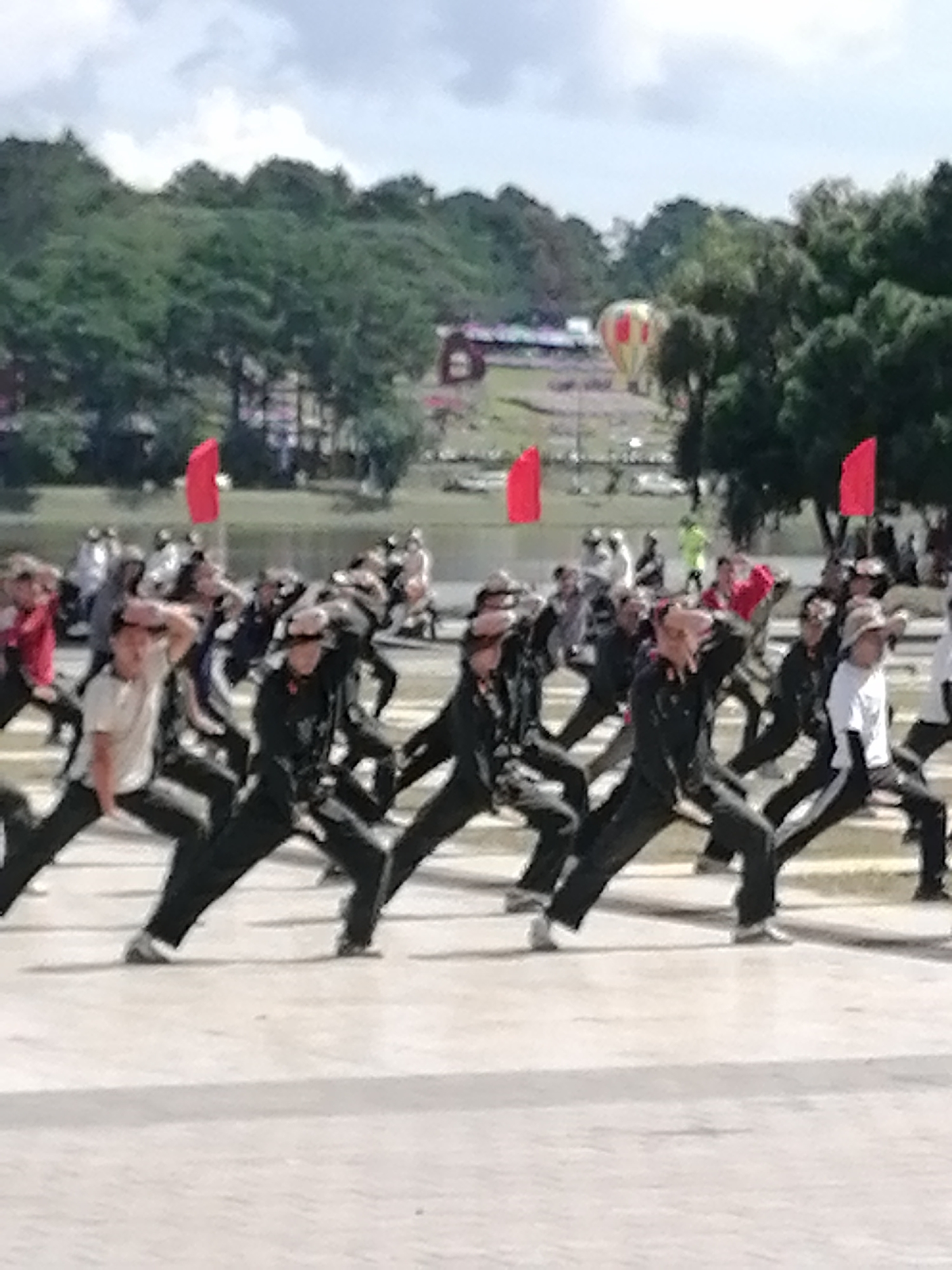 This image captures an outdoor event in Da Lat, Vietnam, likely a synchronized performance or demonstration. In the foreground, a group of individuals, mostly men, are engaged in what appears to be a coordinated physical activity, possibly a martial art or a dance. They are dressed in dark uniforms with some exceptions of lighter colored tops. Their stances suggest a moment of dynamic movement, with many bending their knees and extending their arms.
The background reveals a large body of water, possibly Xuan Huong Lake, a prominent landmark in Da Lat. Beyond the water, a lush green hillside is visible, densely covered with trees. In the distance, above the treeline, two hot air balloons are present, one red and yellow, and another primarily red. Red flags are also visible, strategically placed within the scene, adding a splash of color against the natural backdrop. The sky is overcast with clouds, suggesting a moderate weather condition. The overall impression is one of an organized cultural or sporting event taking place in a scenic urban environment.
