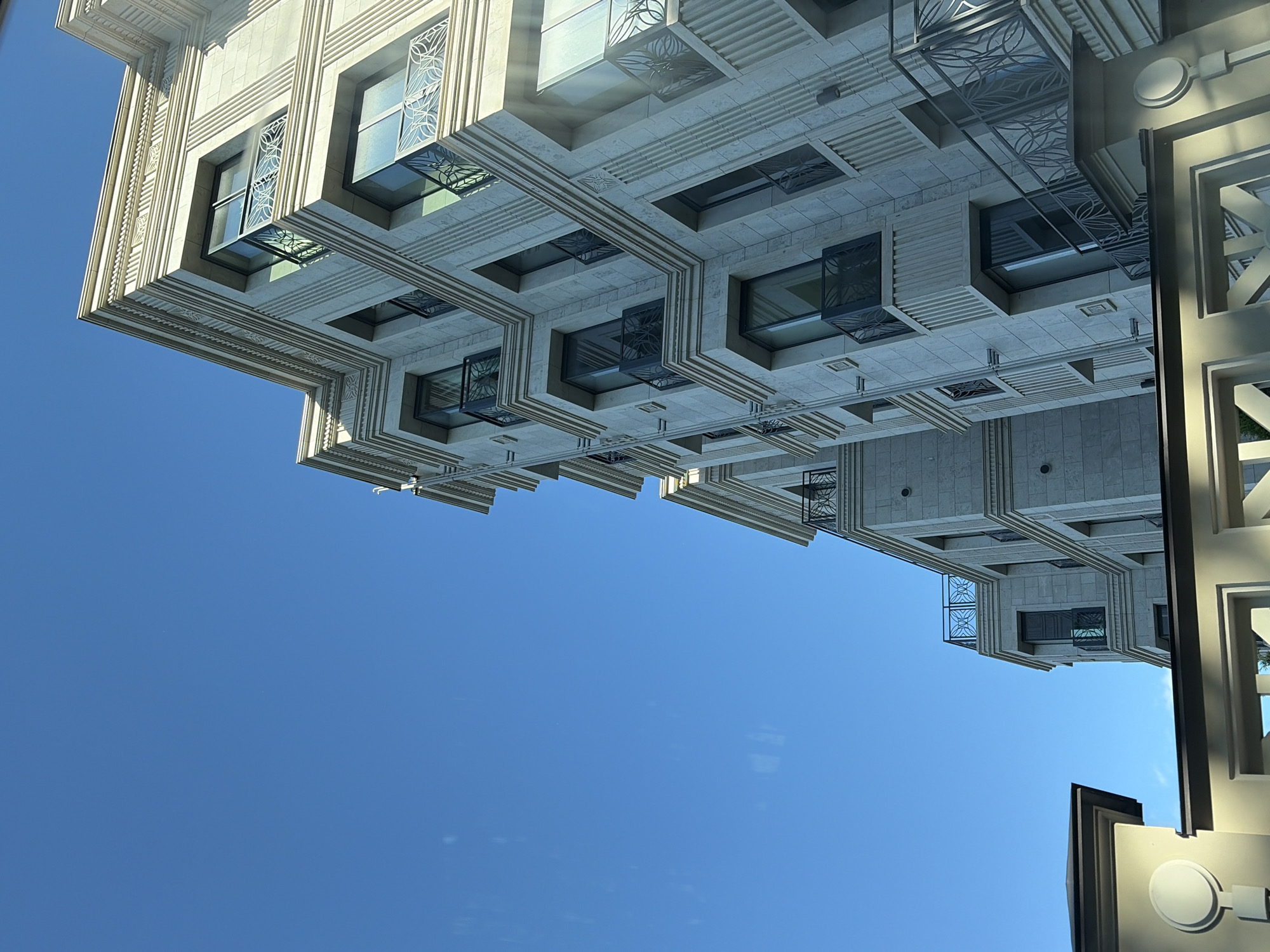 This media file is a photograph taken from a low angle, looking upwards at an ornate, modern building against a clear blue sky. The building features intricate stone detailing, with layered cornices and decorative window grilles. Several windows are visible, some with reflective glass that mirrors the sky. The architectural style suggests a high-end residential or commercial building. The clear blue sky indicates fair weather and likely daytime. The image is oriented such that the building appears to be hanging upside down. There is no visible text in the image. The scene is devoid of people or any specific activity, focusing solely on the architectural features of the building and the sky. The location context of Saint Petersburg, Russia, suggests a potentially European urban setting, though no specific landmarks are identifiable.