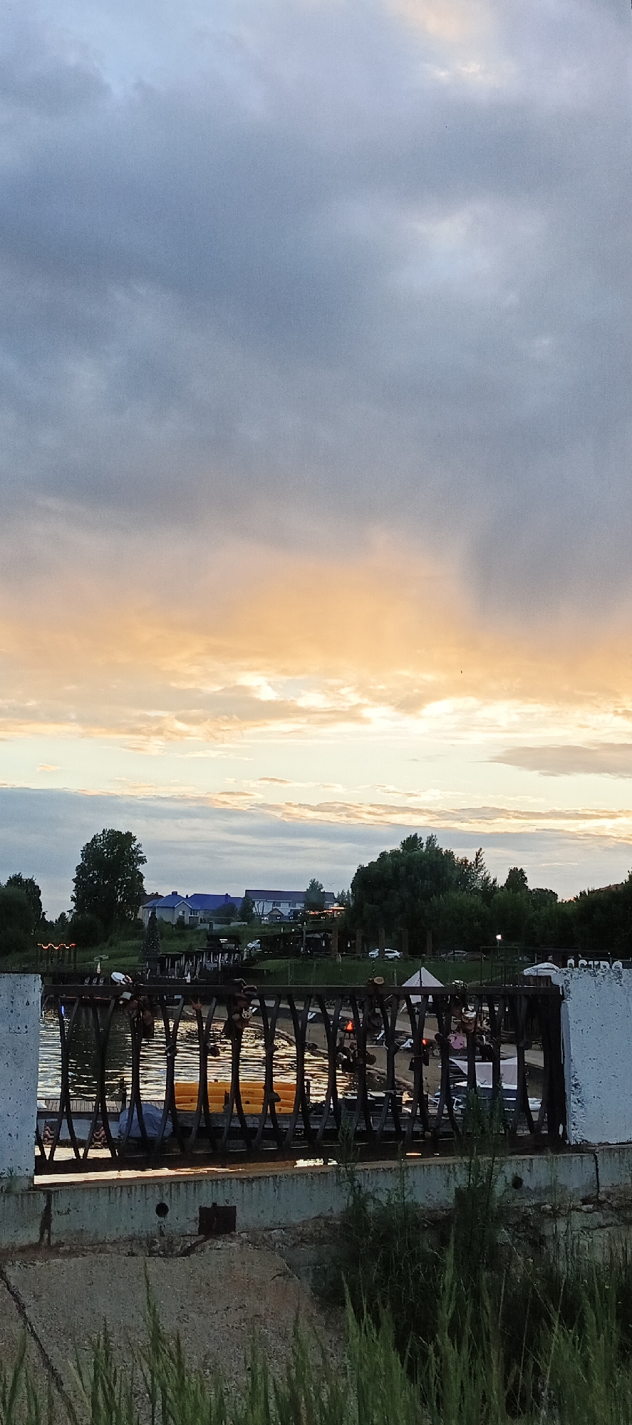 The image depicts a sunset scene over a body of water, likely a river or lake, in Votkinsk, Russia. A dark, ornate metal railing runs horizontally across the foreground, with its reflection visible in the water below. Behind the railing, people are visible on what appears to be a waterfront promenade or embankment. Structures, including buildings with blue roofs and trees, are situated further back on the opposite bank. The sky is filled with dramatic clouds, transitioning from a pale orange and yellow near the horizon to a cooler blue-grey higher up. The overall impression is of a peaceful evening atmosphere.