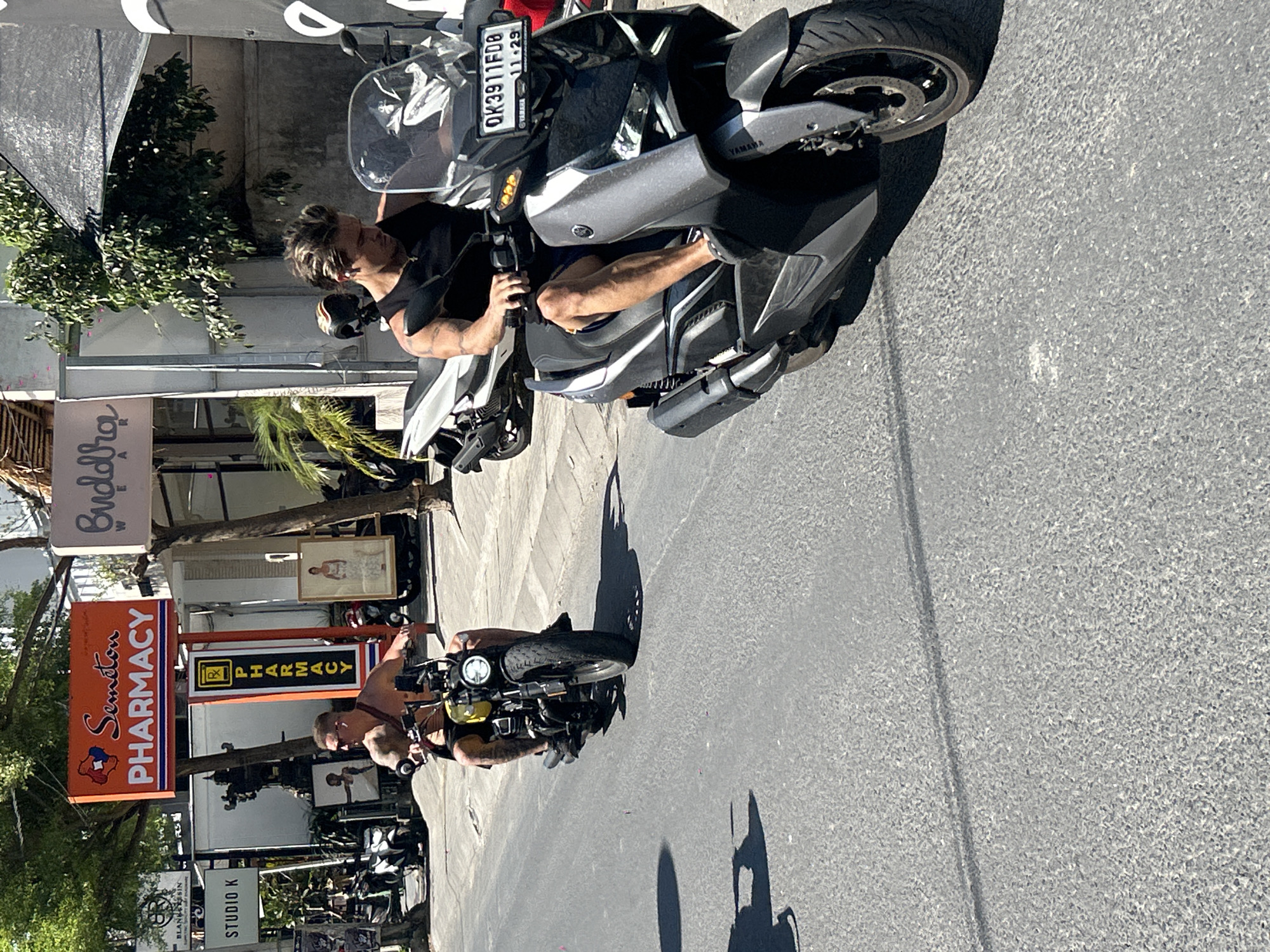 In North Kuta, Indonesia, on a sunlit asphalt road, two men ride motorcycles. In the foreground, a man with blonde-streaked hair, wearing a black tank top and blue shorts, rides a dark gray and silver scooter. His right hand is on the handlebar, and his left arm is bent at the elbow. The scooter has a license plate reading "OK3911FD8". Behind him and to the left, another man, shirtless with tattoos on his arms and chest, rides a yellow and black motorcycle. He is wearing dark shorts and sunglasses. The motorcycle has a round headlight and thick tires. Both men are looking forward, focused on riding. The background features buildings with signs, including "Seminyak Pharmacy" and "Studio K". Lush green trees and palm fronds are visible, suggesting a tropical environment. The lighting indicates it is daytime, with shadows cast on the road.