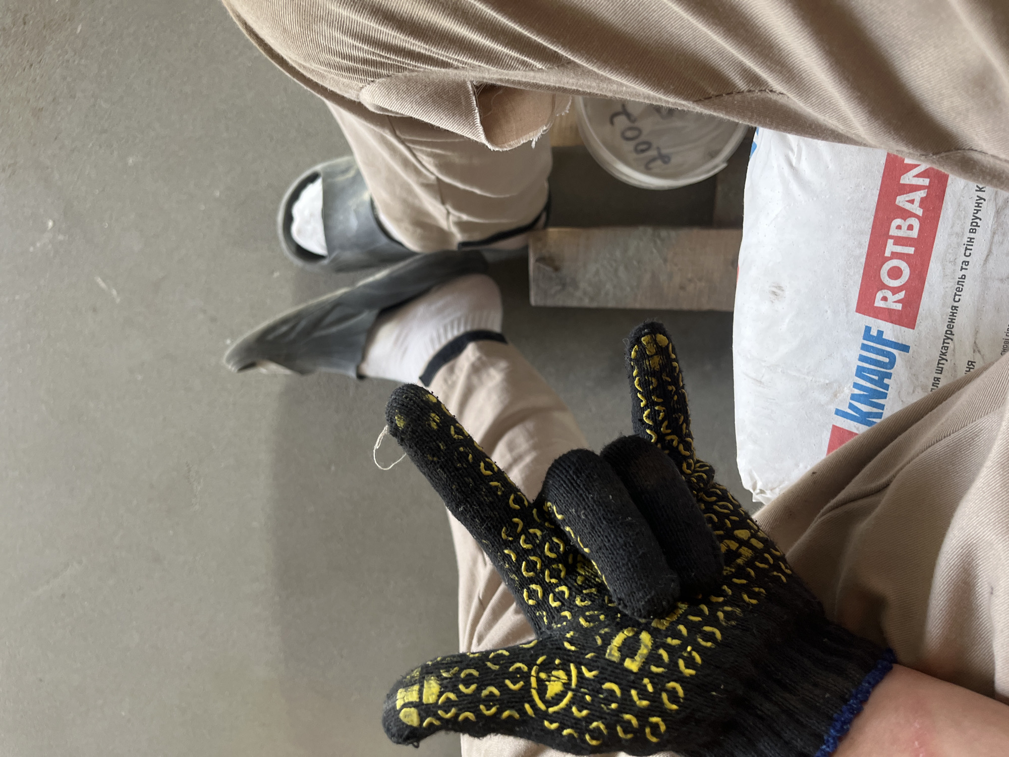 The image shows a close-up view of a person's legs and hand, likely in a construction or workshop setting. The person is wearing tan work pants, white socks, and dark gray slides. They are also wearing a black work glove with yellow patterns on their right hand, and are making a hand gesture. In the background, there is a white bag with "KNAUF ROTBAN" printed on it, and a container with "2002" written on it. The environment appears to be indoors with a concrete floor. The image was taken in Strumivka, Ukraine.