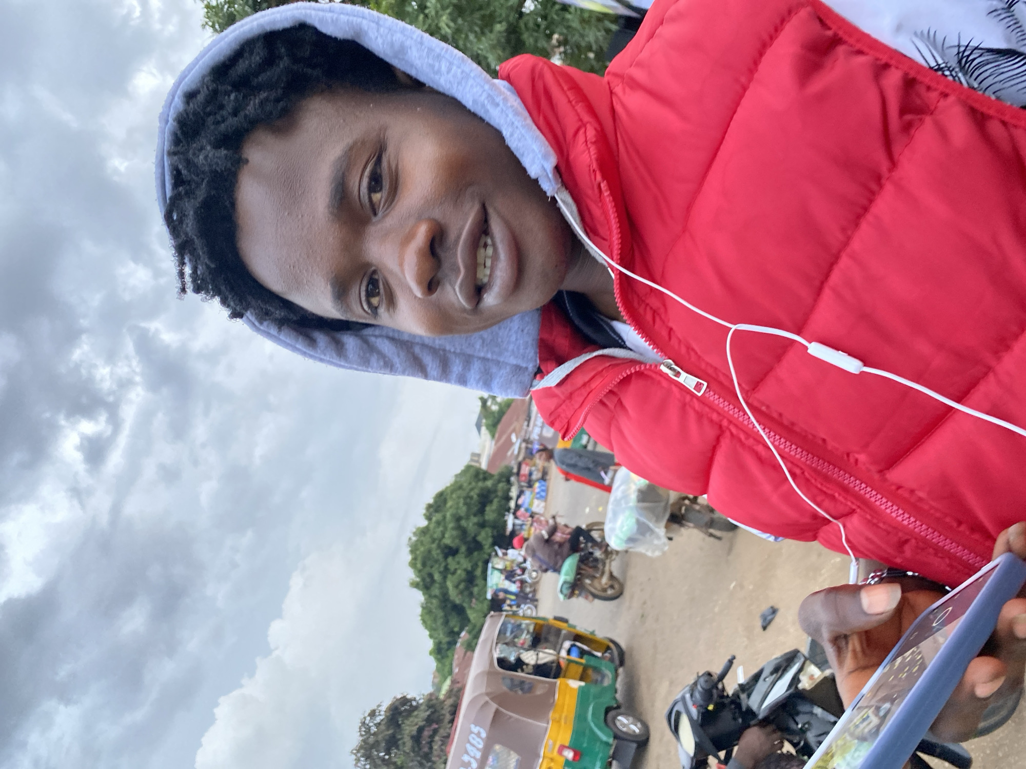 In Bukuru, Nigeria, a young African male, positioned in the foreground and capturing a selfie, stands against a backdrop of a cloudy sky and a bustling street scene. He wears a red puffer vest over a white shirt, with a grey hooded sweatshirt pulled up. White wired earbuds are visible, plugged into his attire. In his left hand, he holds a blue smartphone.
The background reveals a lively street environment. A yellow and green auto-rickshaw, identified by the registration number "3405", is parked to the left. Several motorcycles are also present, with one ridden by a person wearing a red cap and a brown jacket. Other individuals are seen walking and standing near market stalls or buildings. The ground is dirt or gravel, typical of many Nigerian urban areas. The overall atmosphere suggests a typical day in a busy, semi-urban Nigerian locale.